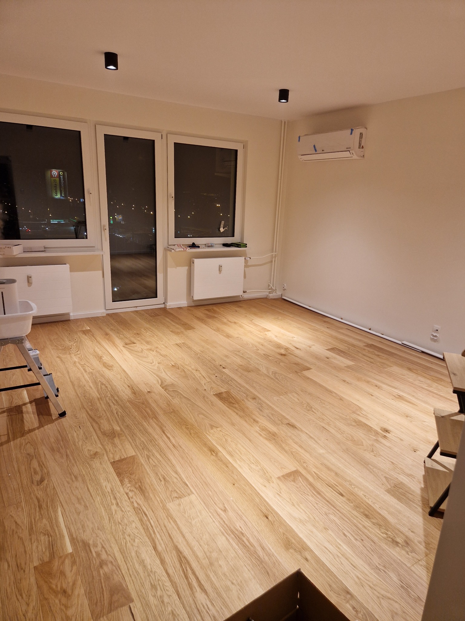 Pusty pokój po remoncie z drewnianą podłogą, grzejnikami pod oknami, klimatyzacją na ścianie i widokiem na nocne miasto za oknem.