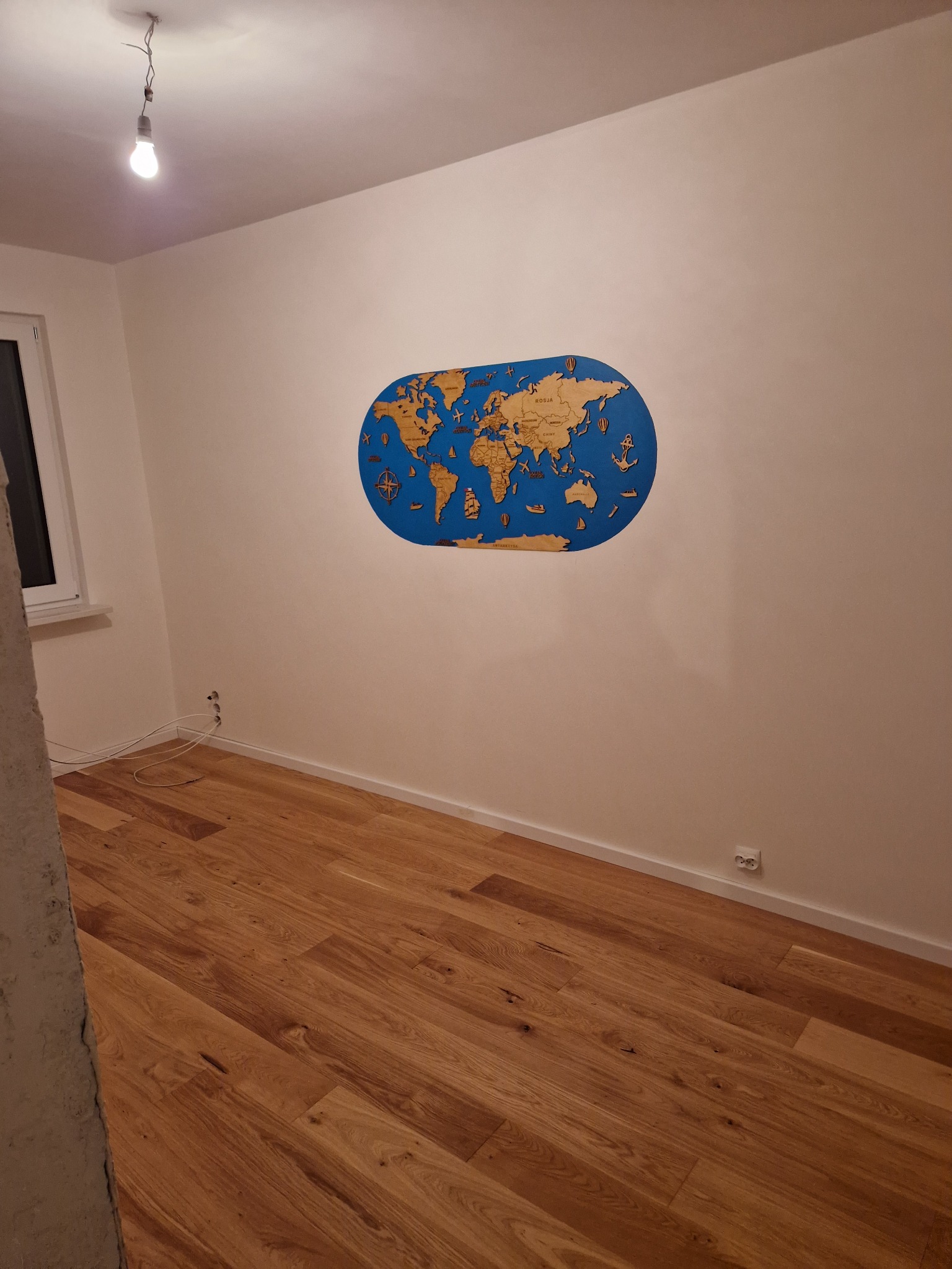 Puste pomieszczenie z drewnianą podłogą i dekoracyjną, drewnianą mapą świata na ścianie. Widoczne gniazdka elektryczne i wisząca żarówka.
