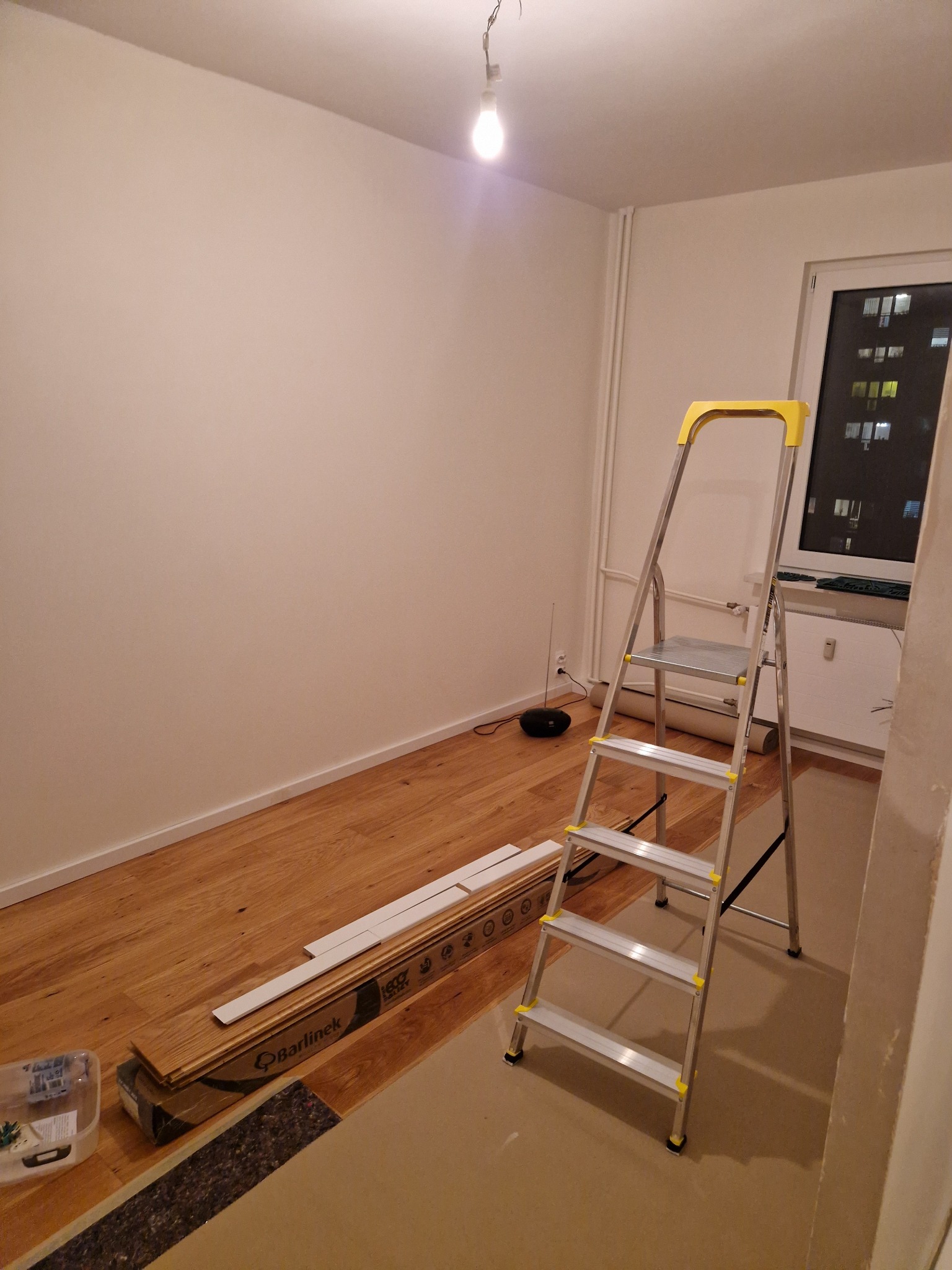 Pokój w trakcie remontu: ułożona drewniana podłoga Barlinek, drabina aluminiowa z żółtymi elementami, białe ściany, surowe oświetlenie żarówką.