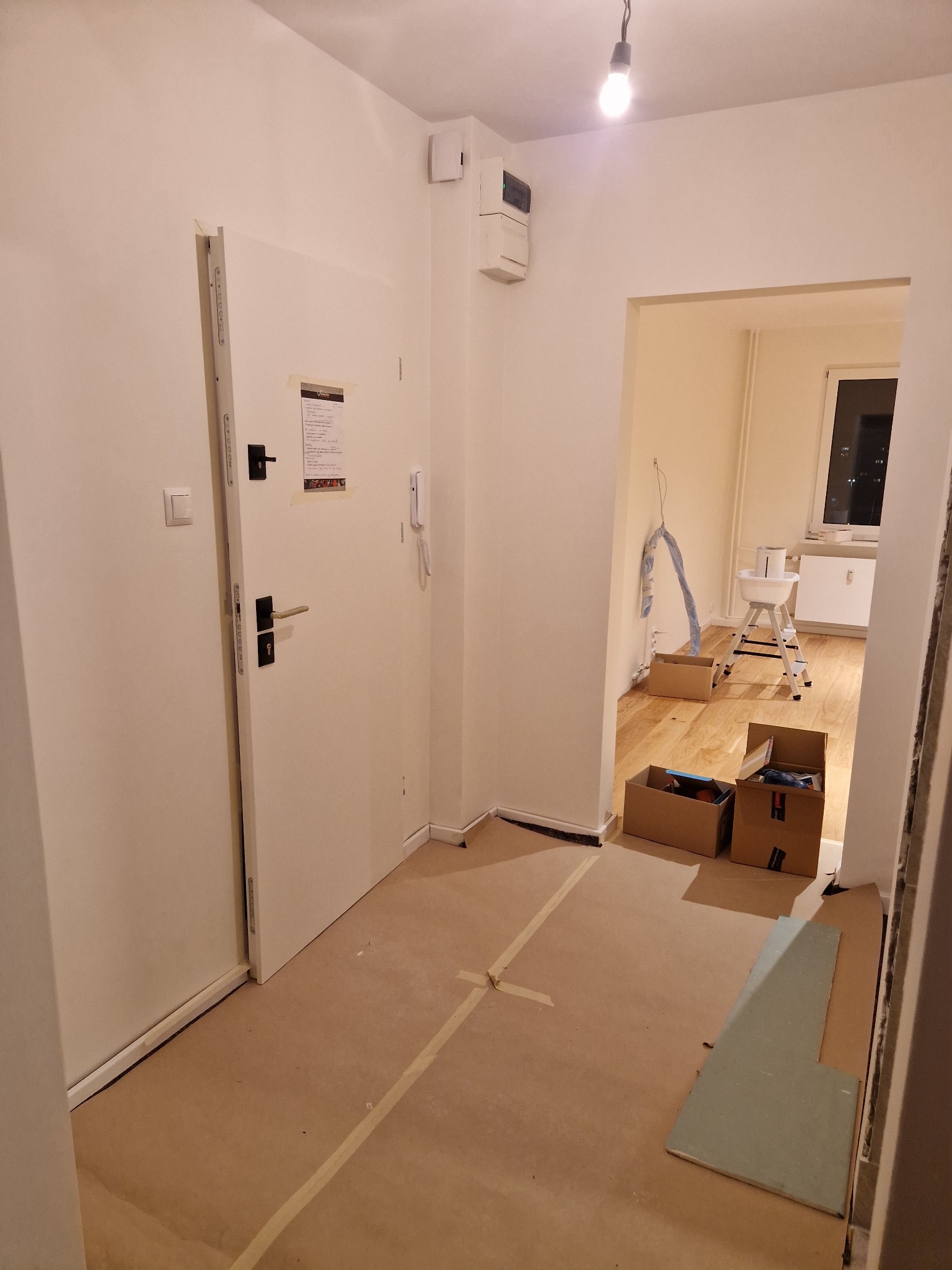 Przedpokój w trakcie zabezpieczania podłogi kartonem i taśmą przed pracami wykończeniowymi, widoczne otwarte kartony i drabinka malarska w tle.