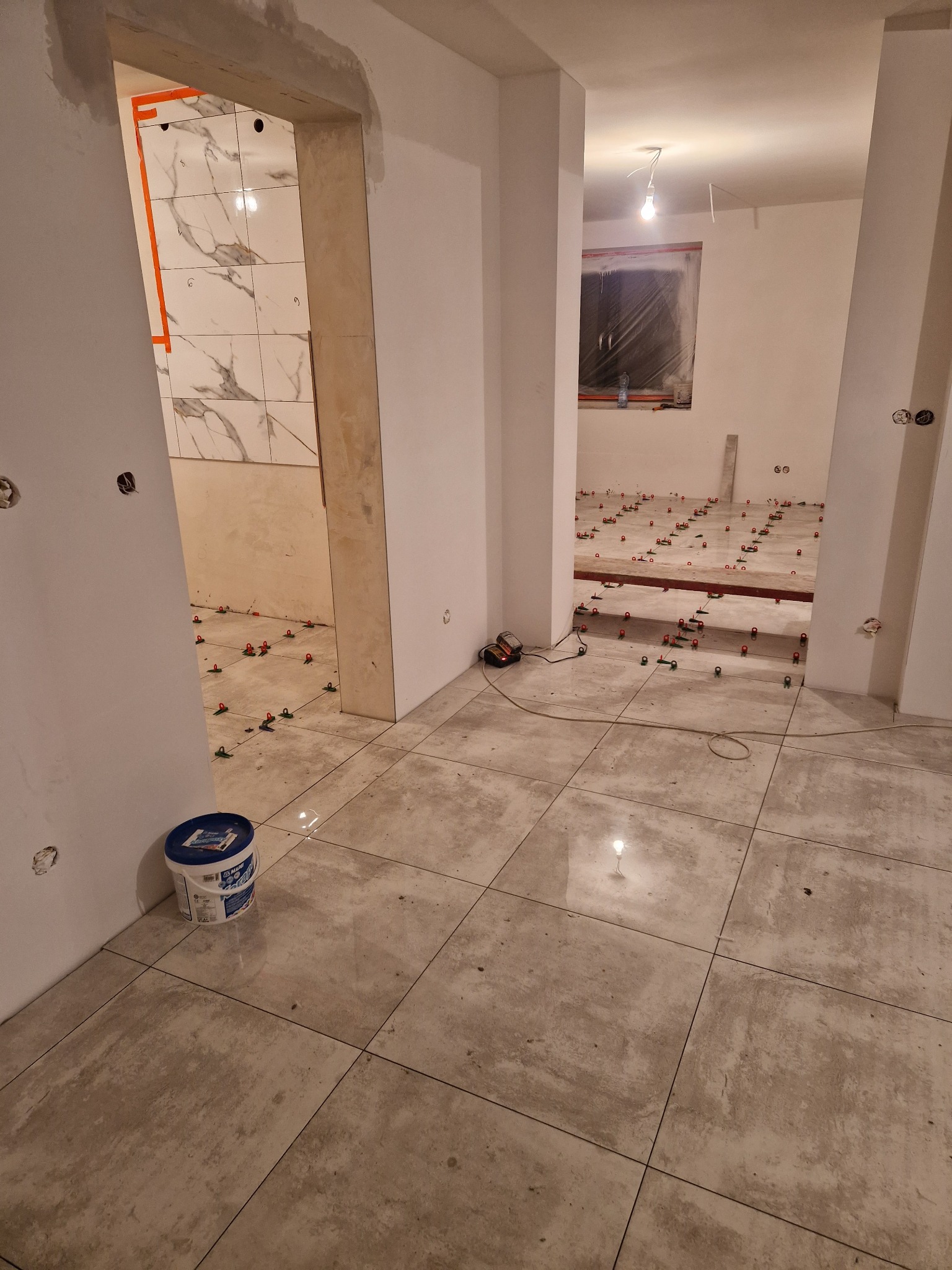 Wnętrze w trakcie układania płytek podłogowych, widoczne krzyżyki dystansowe i wiadro z klejem przy ścianie, w oddali łazienka z częściowo wyłożonymi marmurowymi płytkami.