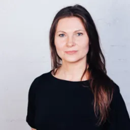 Portret kobiety o jasnych oczach, w czarnej bluzce, na tle białej ceglanej ściany z widocznymi pionowymi rurami po prawej stronie kadru. Uśmiech na twarzy.