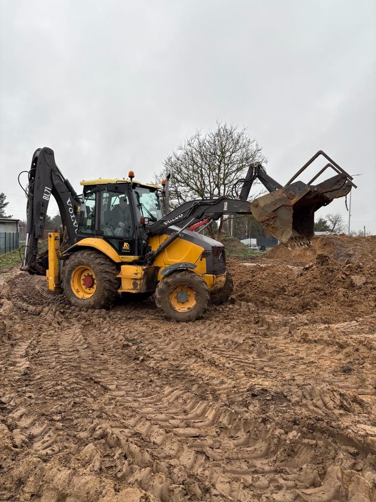 Żółta koparko-ładowarka Volvo w trakcie pracy na placu budowy, z widocznymi śladami opon w błotnistym terenie, w tle drzewa i ogrodzenie.