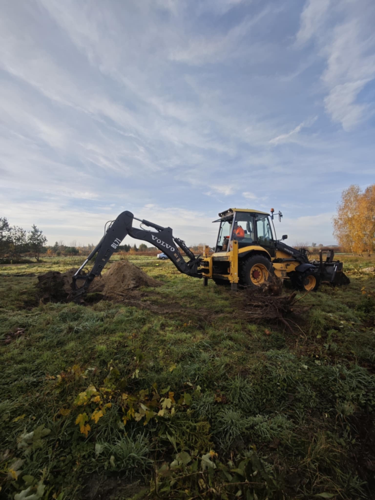 Żółta koparko-ładowarka Volvo BL71 kopie ziemię na zewnątrz, w tle widoczne drzewa i błękitne niebo z chmurami.