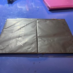 Komputeron - Serwis komputerowy - Dwie czarne, prostokątne baterie litowo-jonowe leżące obok siebie na niebieskim, lekko zabrudzonym stole. Na jednej baterii widoczne napisy informacyjne i ostrzegawcze w różnych językach, w tym...