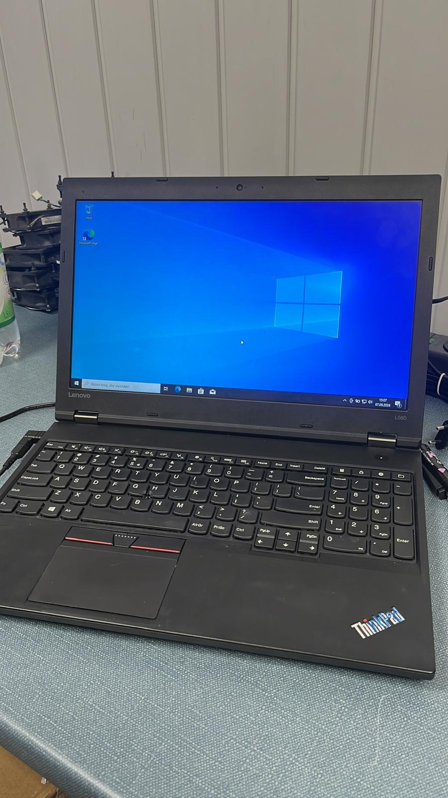 Czarny laptop Lenovo ThinkPad L560 z włączonym ekranem Windows, widoczne logo ThinkPad w prawym dolnym rogu, klawiatura QWERTY i touchpad, w tle części komputerowe i butelka z płynem.
