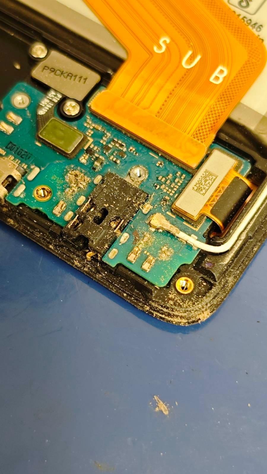 Zbliżenie na zanieczyszczoną płytę główną smartfona z widocznymi elementami elektronicznymi, w tym złączem słuchawkowym i pomarańczową taśmą z napisem 'SUB'.