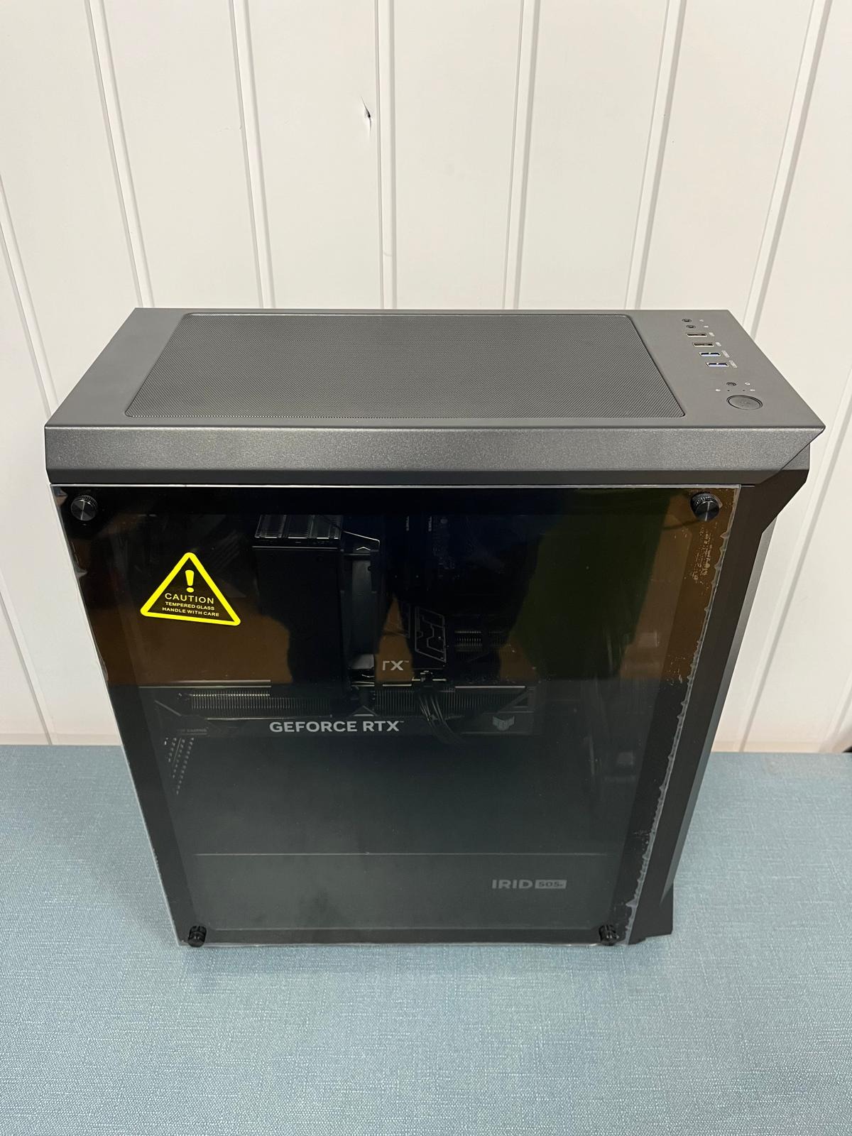 Komputer stacjonarny w czarnej obudowie z widoczną kartą graficzną GeForce RTX i naklejką ostrzegawczą na szklanym panelu bocznym.