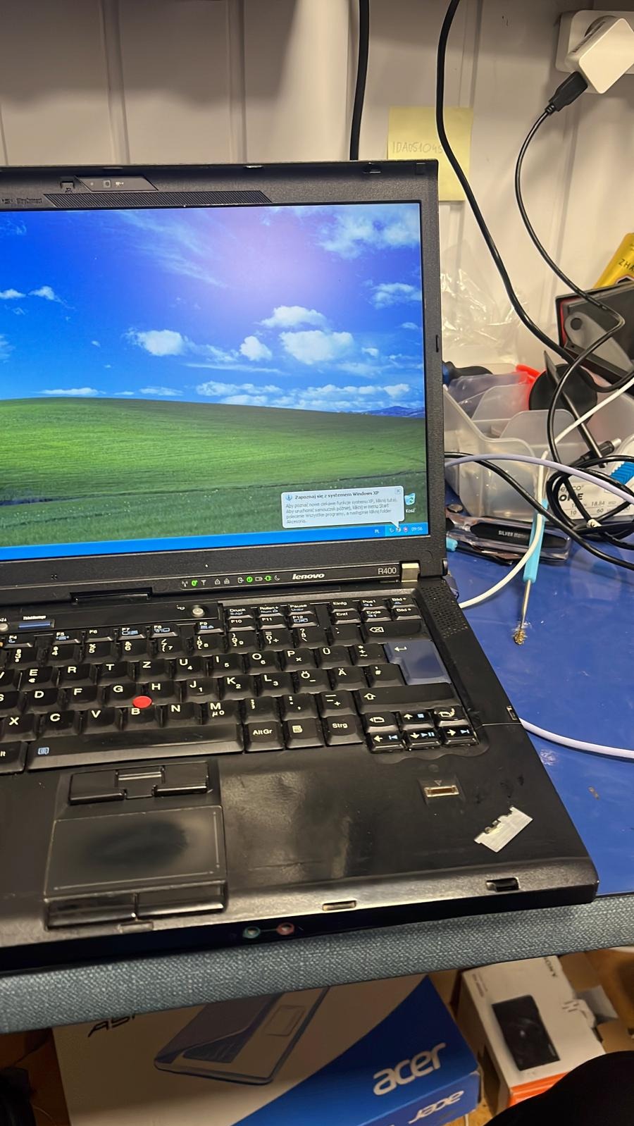 Używany laptop Lenovo R400 z systemem Windows XP, widoczny pulpit z domyślną tapetą, na biurku z narzędziami i przewodami.