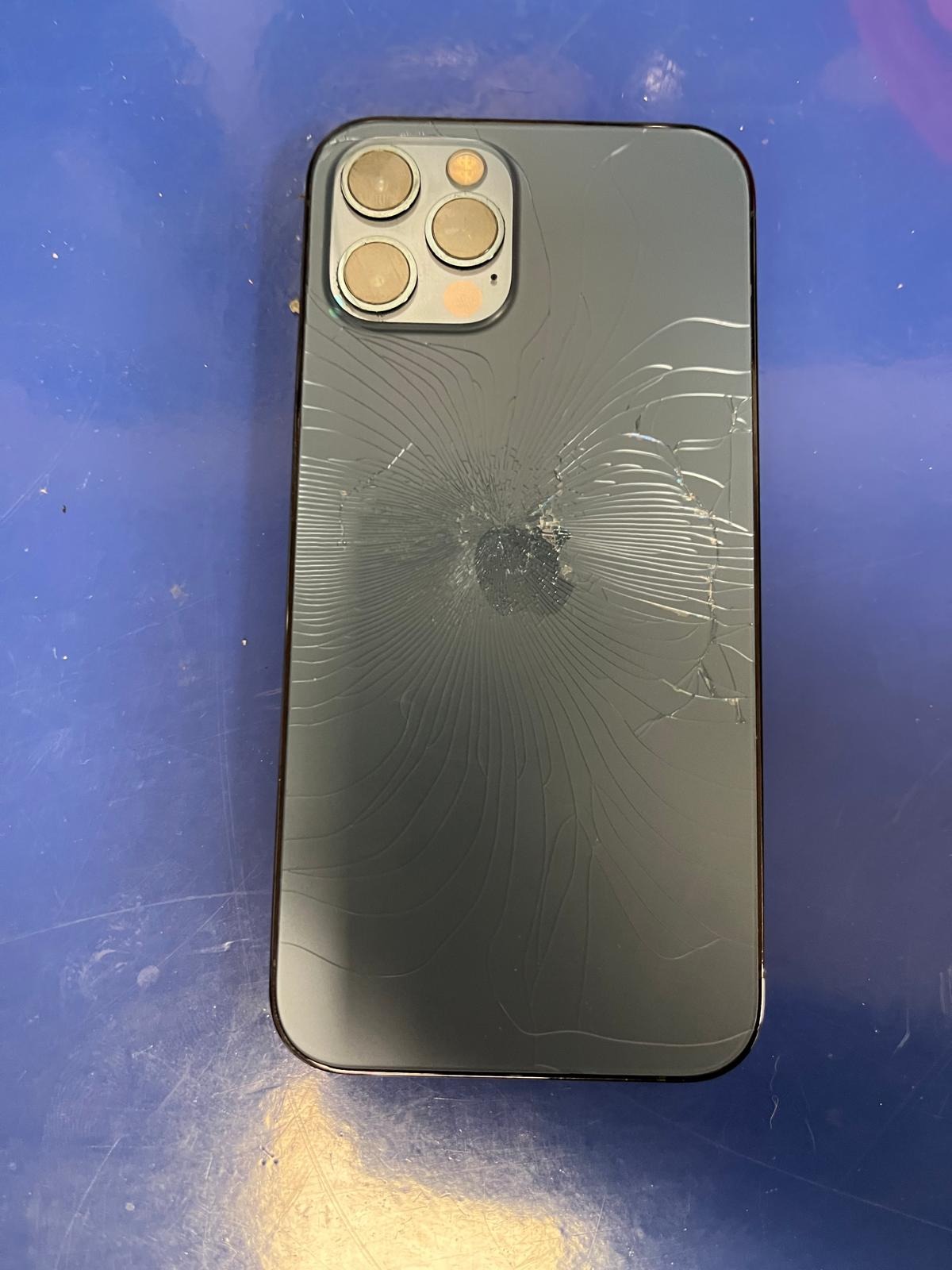 Smartfon z mocno uszkodzoną, pękniętą obudową na niebieskim tle, widoczne pęknięcia rozchodzące się promieniście od punktu uderzenia.