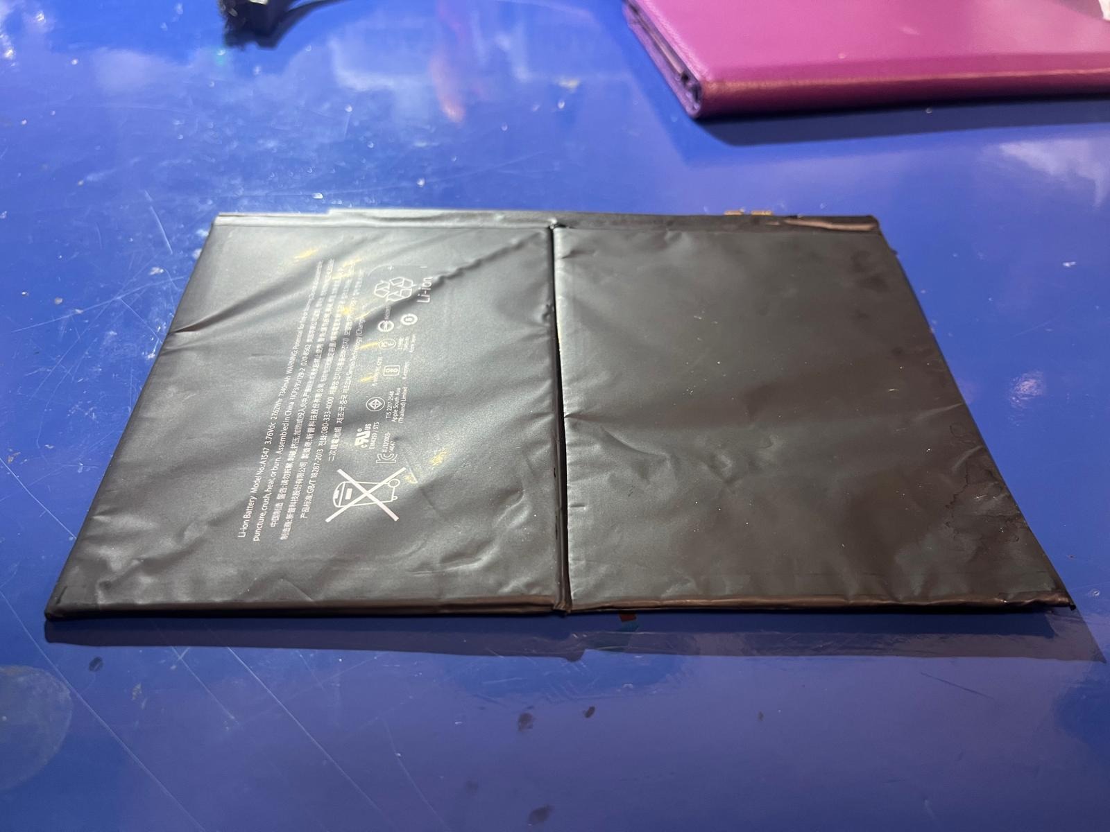 Dwie czarne, prostokątne baterie litowo-jonowe leżące obok siebie na niebieskim, lekko zabrudzonym stole. Na jednej baterii widoczne napisy informacyjne i ostrzegawcze w różnych językach, w tym...