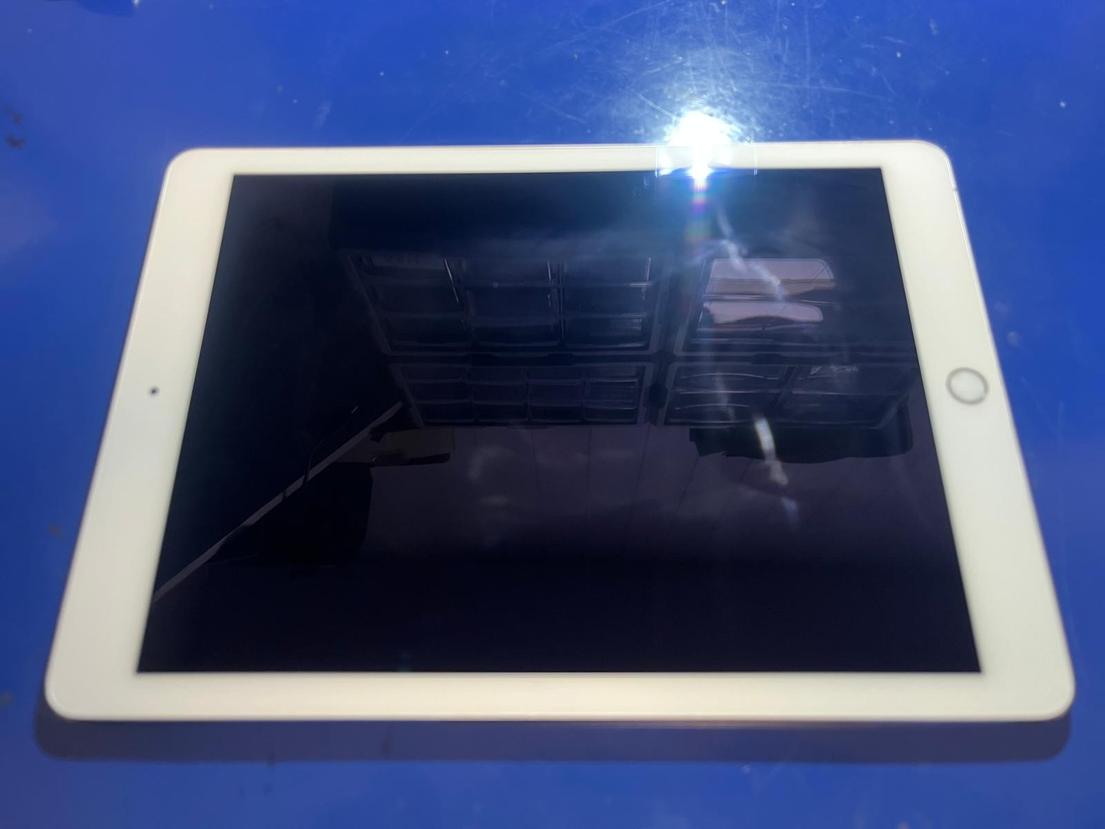 Tablet z wyłączonym ekranem leżący na niebieskim tle, na ekranie widoczne odbicie szafki z przegródkami.