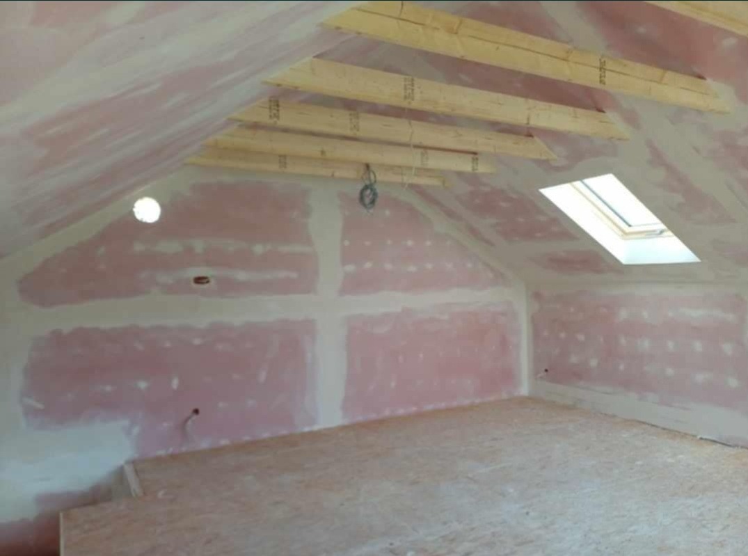Poddasze w trakcie przygotowania do malowania: ściany pokryte gładzią gipsową i gruntem, widoczne drewniane elementy konstrukcji dachu i okno dachowe.