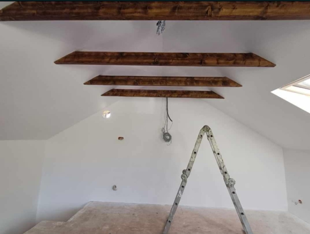 Białe wnętrze poddasza z drewnianymi belkami stropowymi, widoczna drabina malarska i niedokończona podłoga.