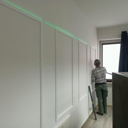 Henzbud - Pracownik montuje panele dekoracyjne na ścianie, używając poziomicy laserowej do precyzyjnego wyznaczenia linii, widoczne narzędzia i fragment szafki.