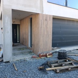 Henzbud - Elewacja domu w trakcie wykończenia z drewnopodobną okładziną, widoczne materiały budowlane i narzędzia na żwirowym podłożu.