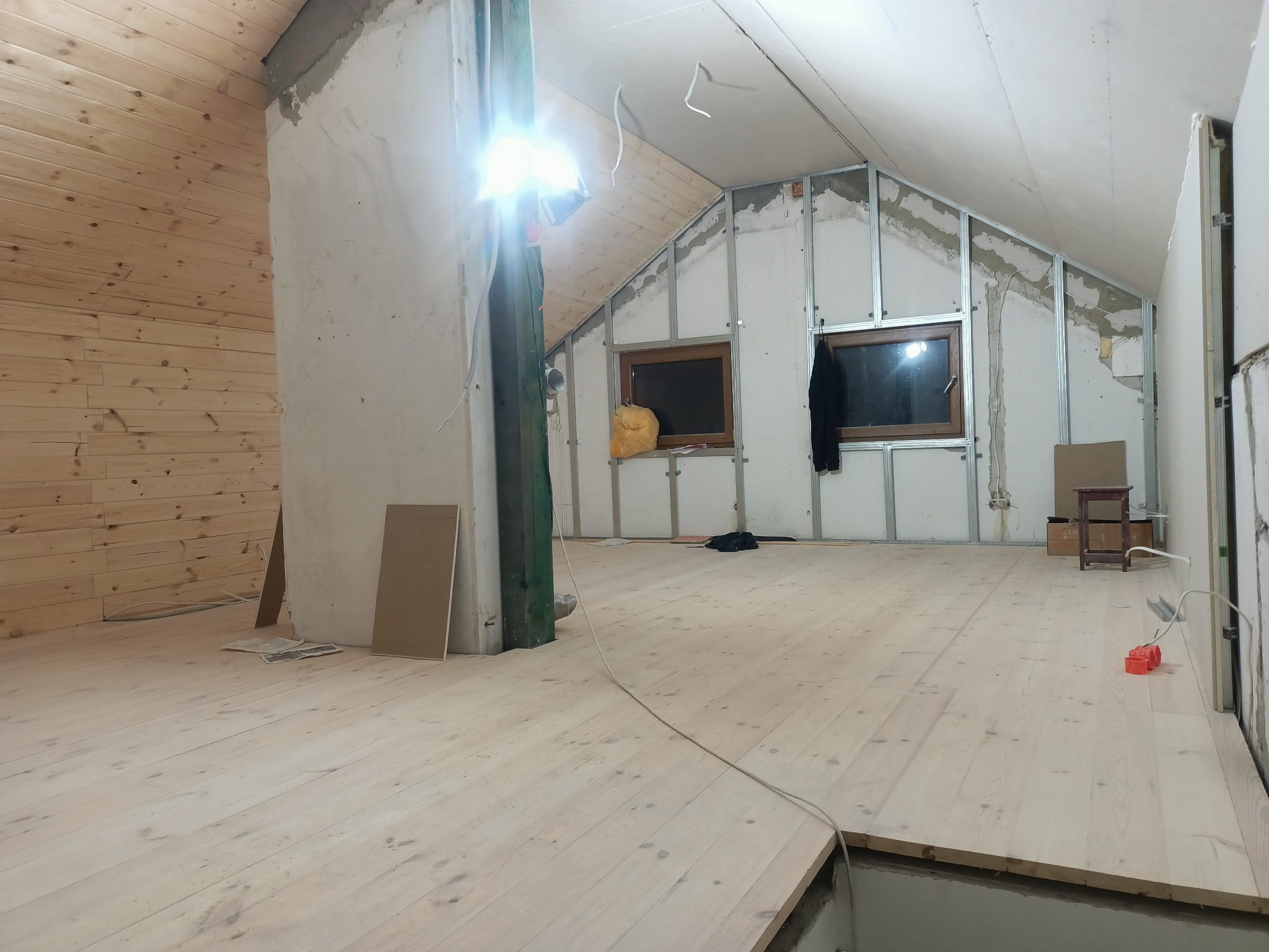 Poddasze w trakcie adaptacji: drewniana boazeria na jednej ścianie, szkielet z profili i płyty gipsowo-kartonowej na pozostałych, drewniana podłoga, okna dachowe w drewnianych ramach, widoczne...