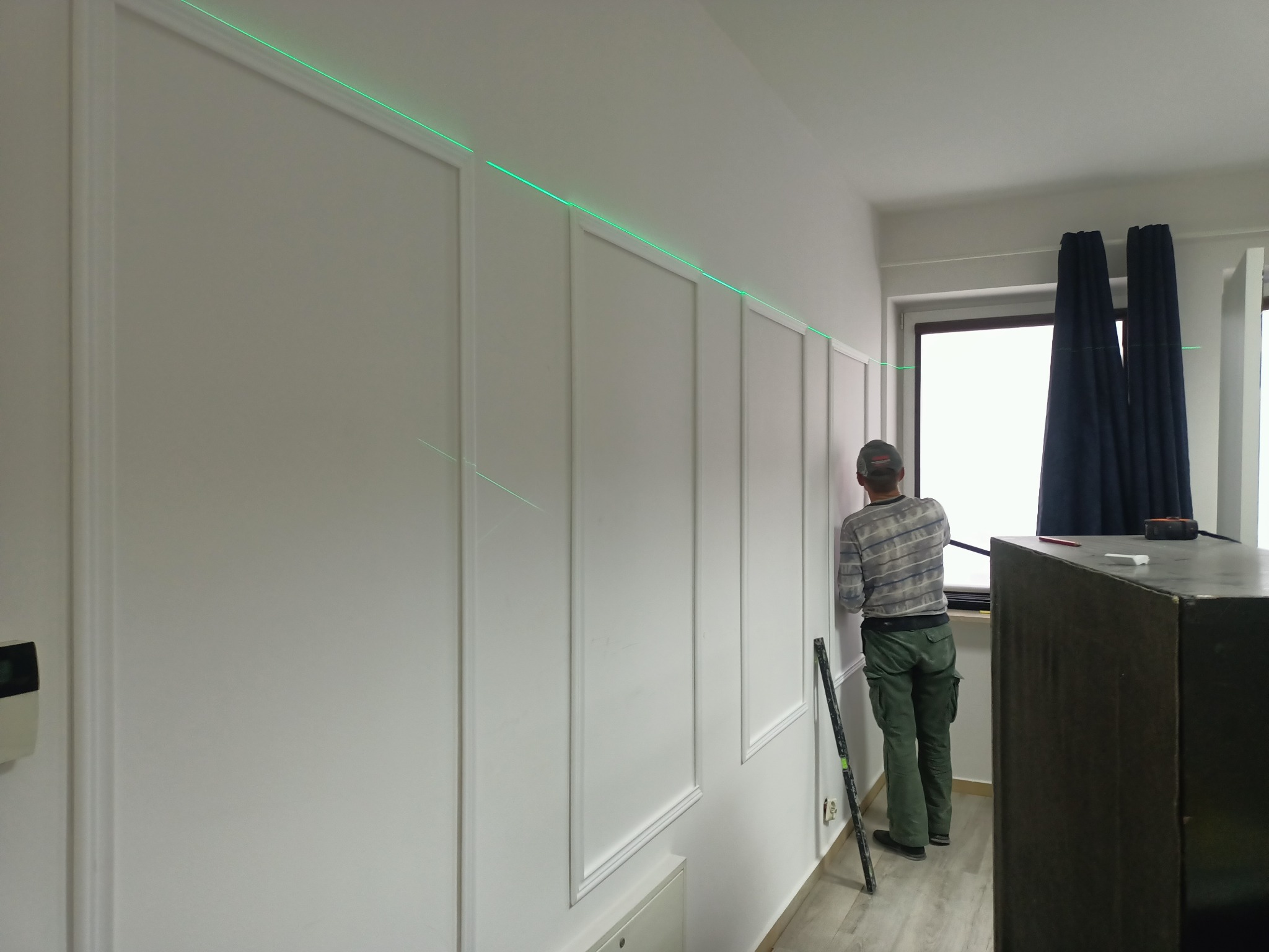 Pracownik montuje panele dekoracyjne na ścianie, używając poziomicy laserowej do precyzyjnego wyznaczenia linii, widoczne narzędzia i fragment szafki.