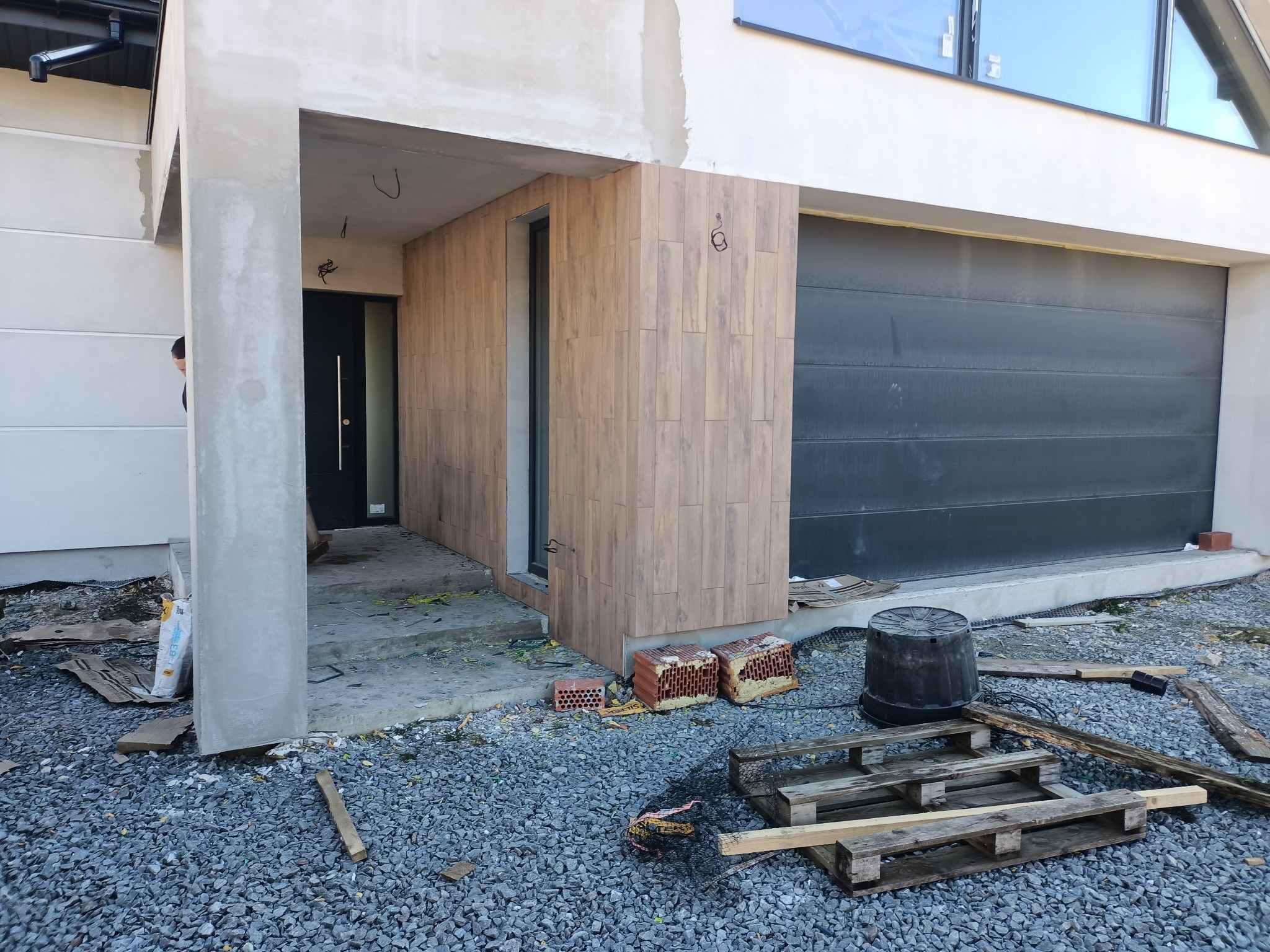 Elewacja domu w trakcie wykończenia z drewnopodobną okładziną, widoczne materiały budowlane i narzędzia na żwirowym podłożu.