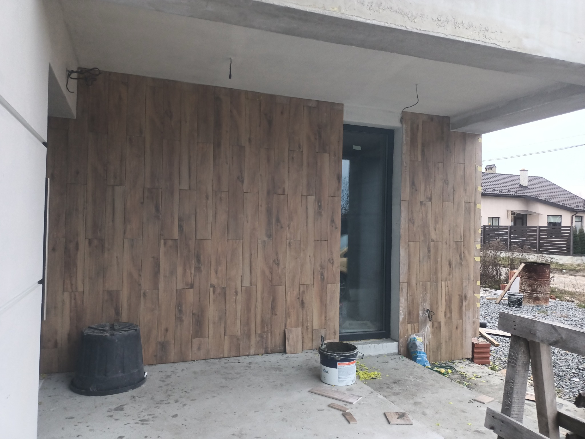 Montaż brązowych płytek imitujących drewno na elewacji budynku w trakcie realizacji, widoczny kubeł z klejem i resztki zaprawy.