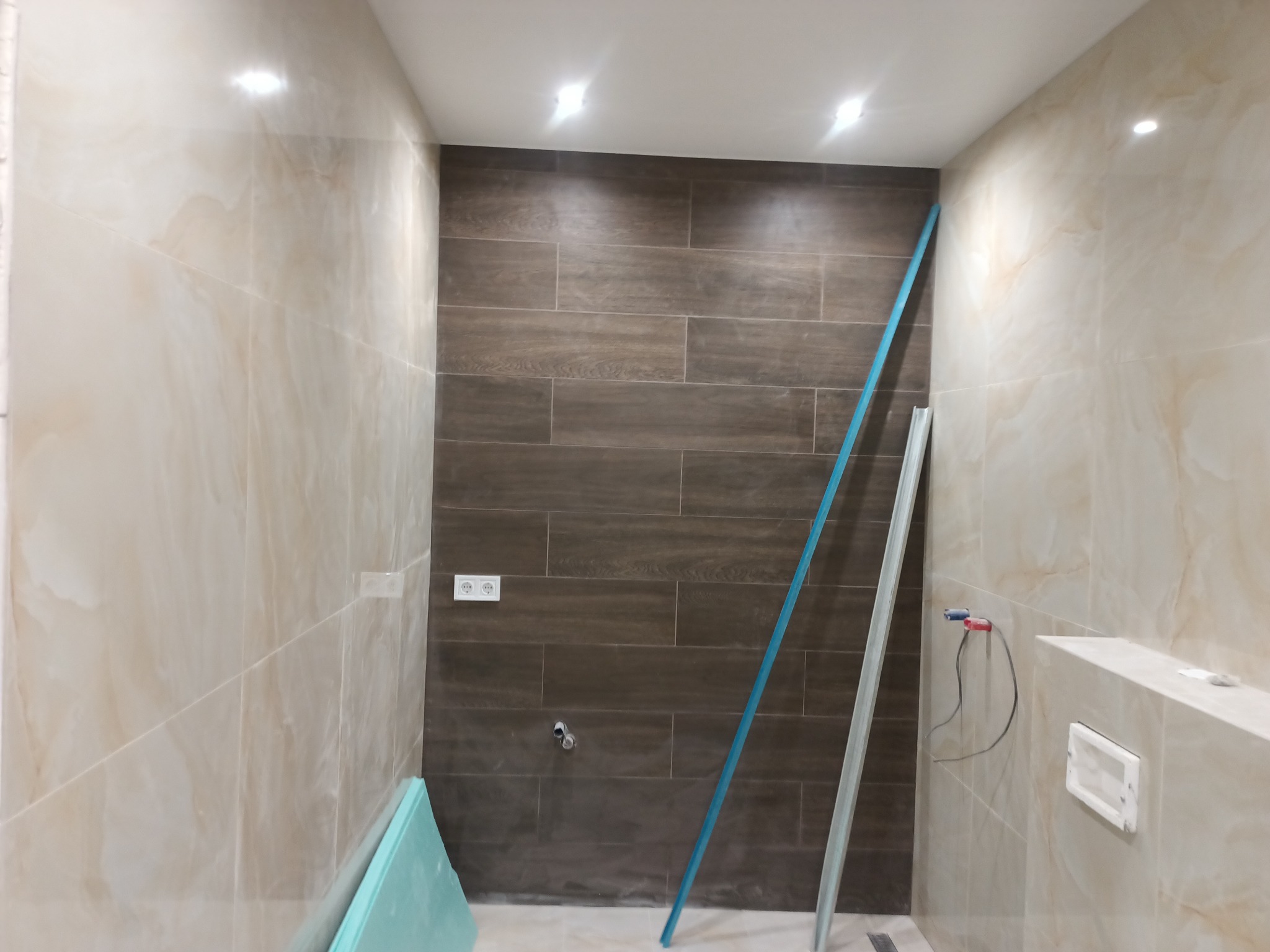 Wnętrze łazienki w trakcie remontu, z częściowo położonymi płytkami imitującymi drewno na jednej ścianie i marmurkowymi płytkami na pozostałych, widoczne profile wykończeniowe i materiały budowlane.