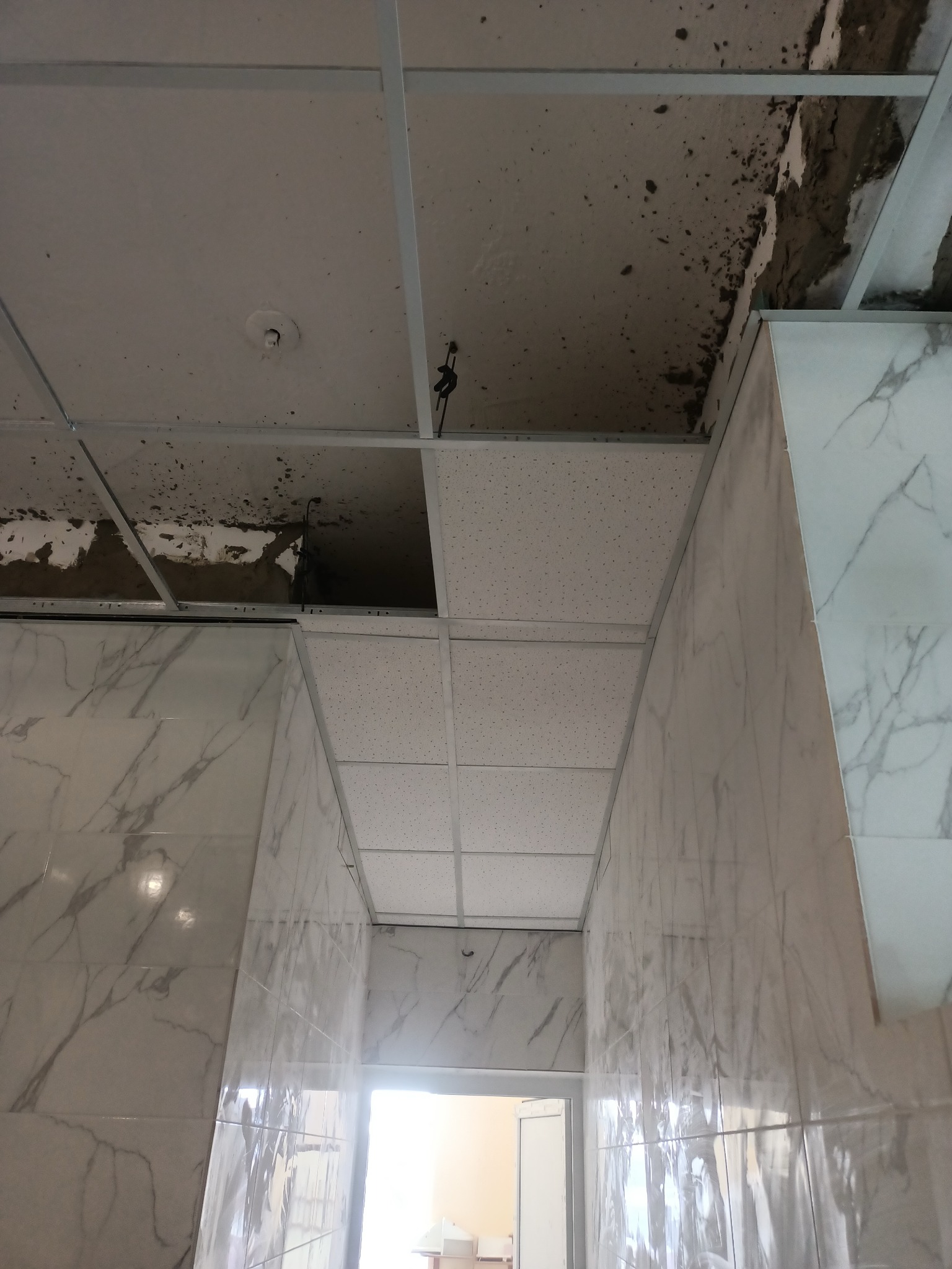 Montaż sufitu podwieszanego z płyt kartonowo-gipsowych w pomieszczeniu z marmurowymi płytkami ściennymi, widoczne niedokończone fragmenty sufitu i instalacje.