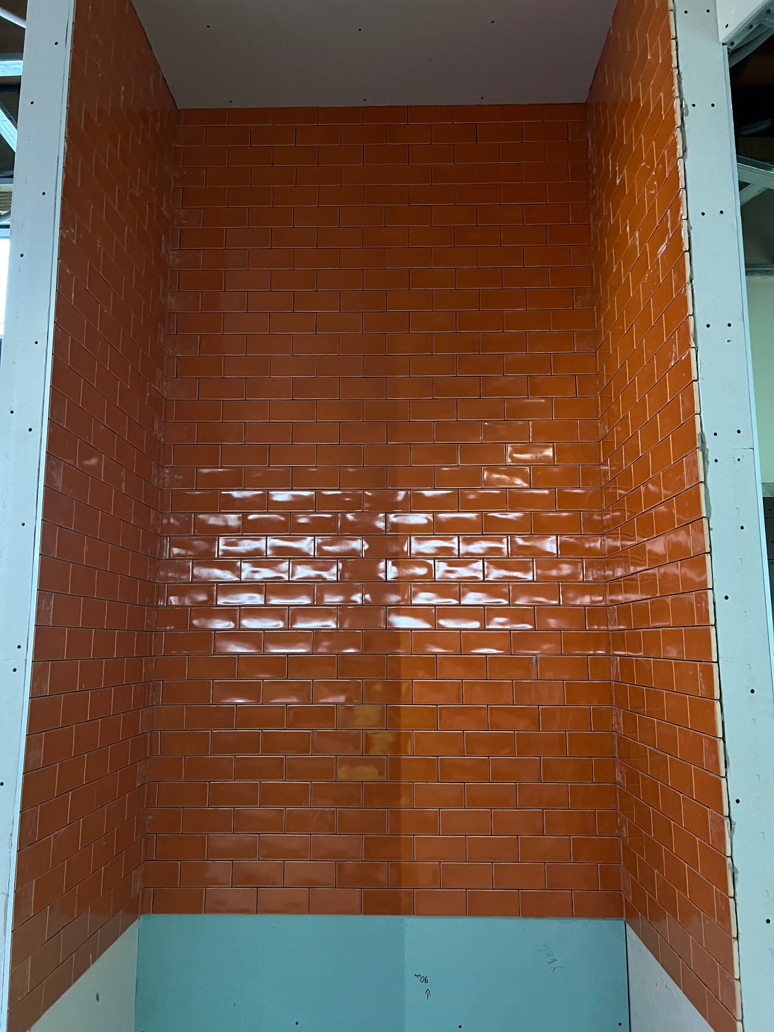 Pomarańczowe, błyszczące płytki ułożone w cegiełkę na ścianie wnęki, obudowane płytą gipsową. Widoczne metalowe profile konstrukcyjne.