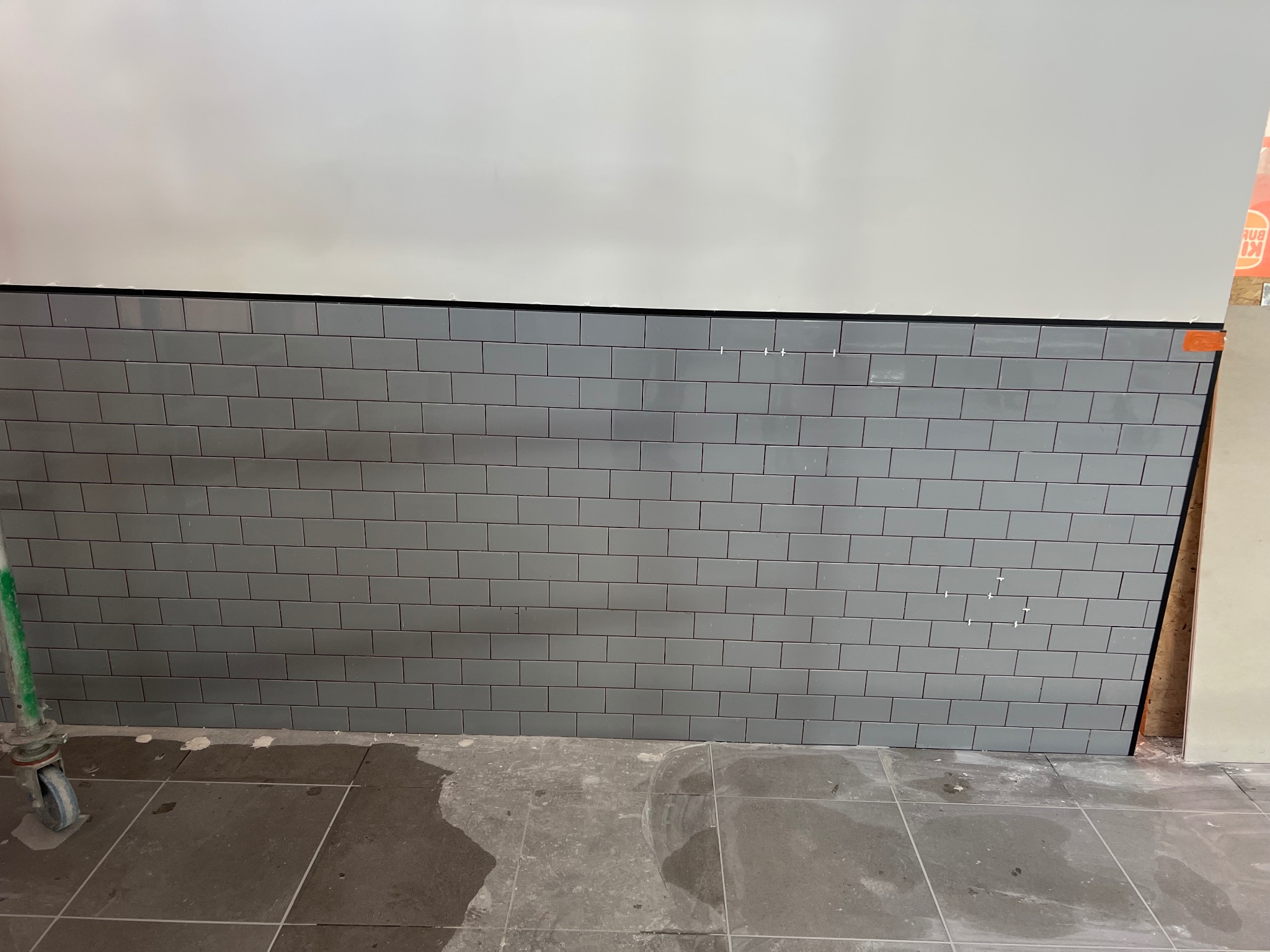 Ściana wyłożona szarymi, prostokątnymi płytkami w stylu metra, z czarną fugą. Widoczne pozostałości po montażu i fragment podłogi z płytek.