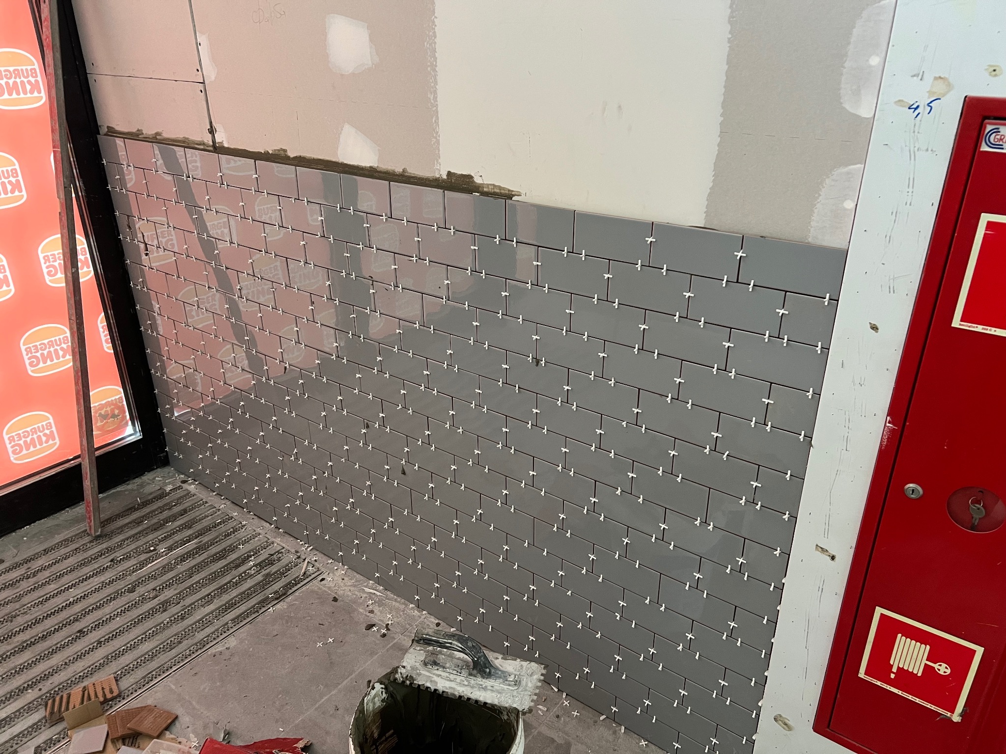 Układanie szarych płytek w restauracji, widoczne krzyżyki dystansowe, wiadro z zaprawą i fragment czerwonej szafki hydrantowej.