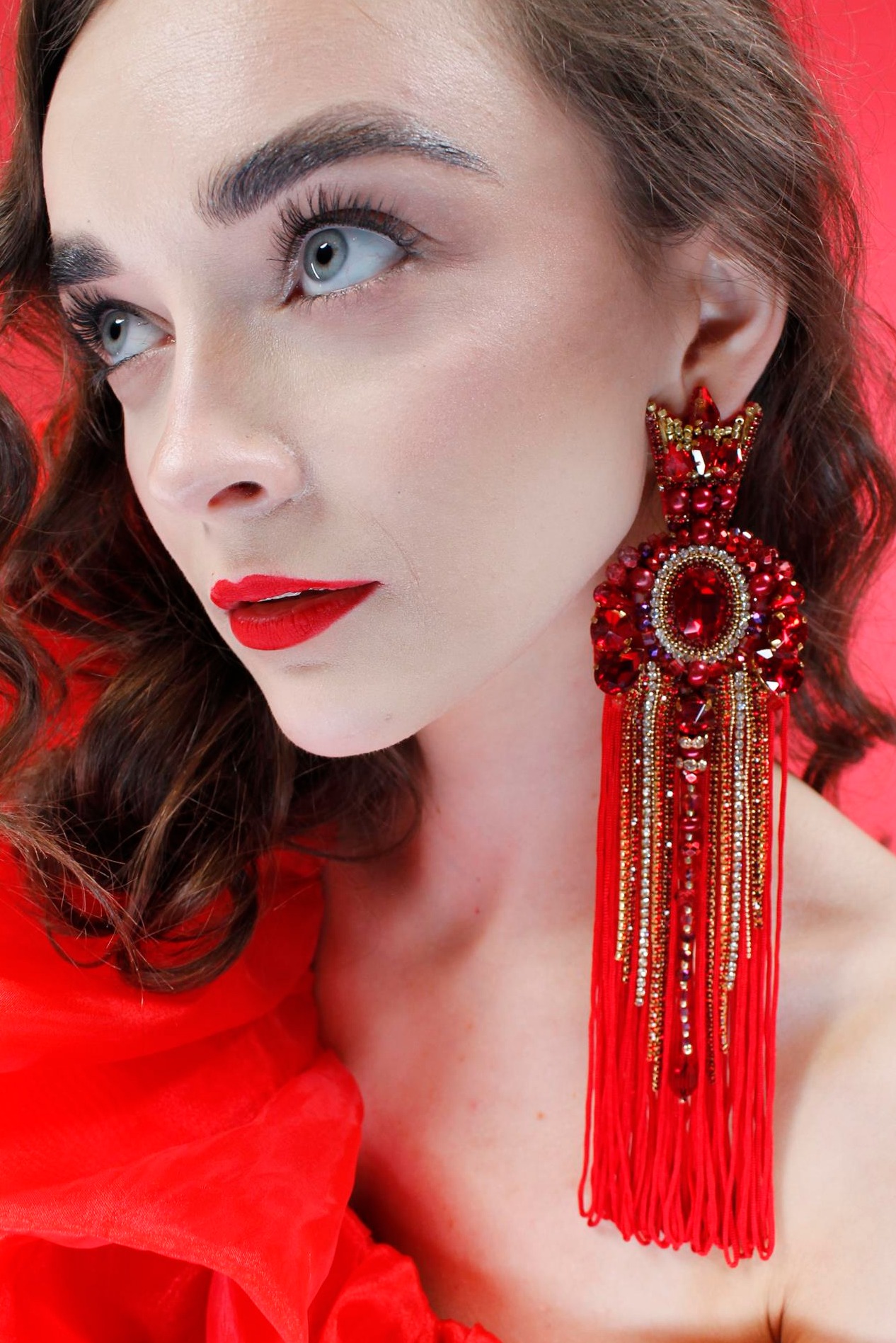 Portret kobiety z intensywnym makijażem ust i dużym, ozdobnym, czerwonym kolczykiem z koralików i frędzli, na czerwonym tle.