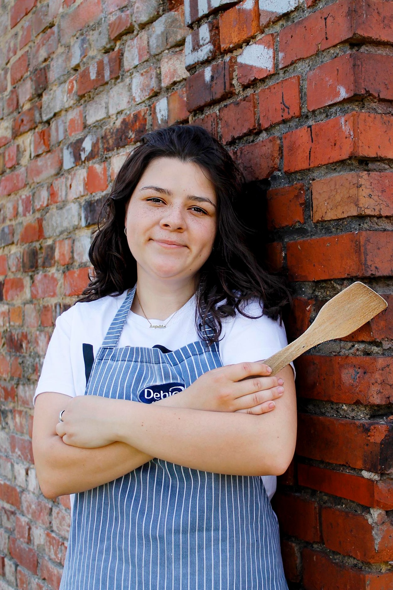 Uśmiechnięta kobieta w niebiesko-białym fartuchu w paski, oparta o ceglaną ścianę, trzyma drewnianą łopatkę do mieszania.