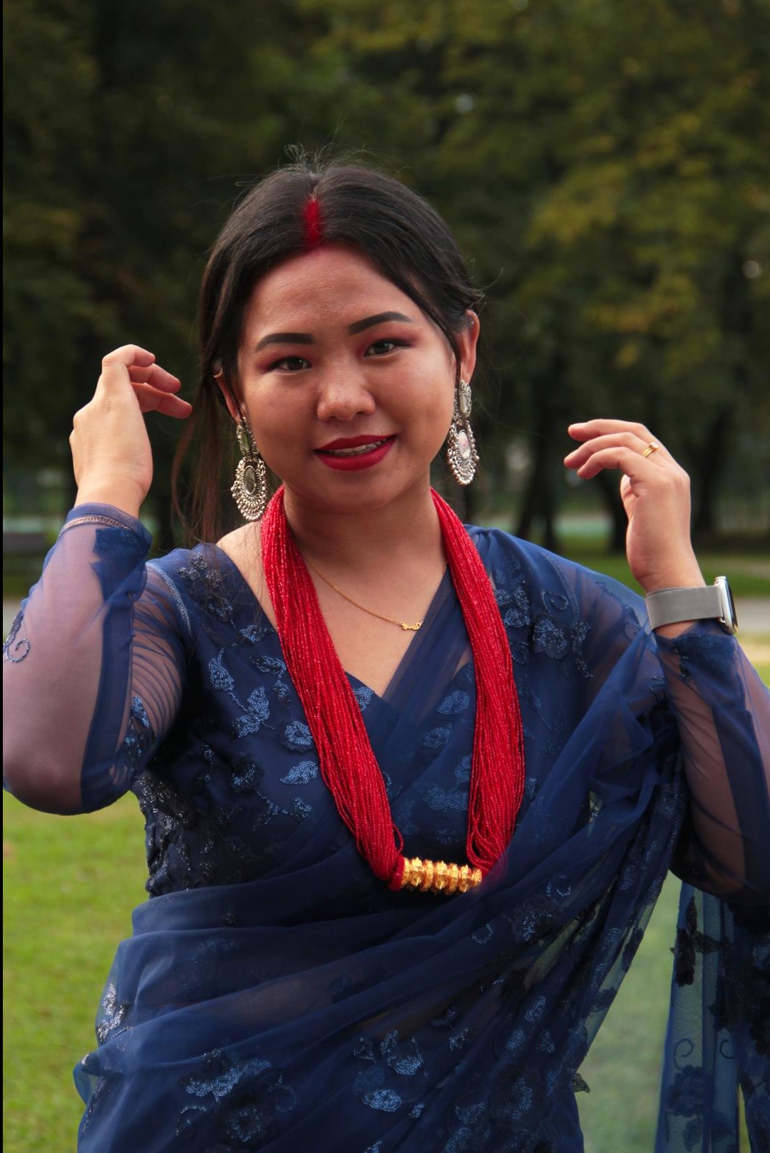 Uśmiechnięta kobieta w niebieskim sari i tradycyjnej biżuterii pozuje na tle zieleni.