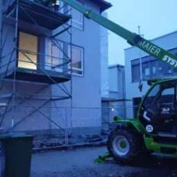 Montaż okna na rusztowaniu przy użyciu podnośnika teleskopowego Maier System na tle budynku w trakcie budowy.