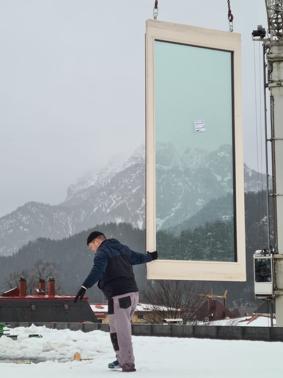 Montaż dużego okna na dachu budynku w zimowej scenerii; pracownik w czapce i rękawicach stabilizuje okno zawieszone na dźwigu na tle gór i zabudowań.