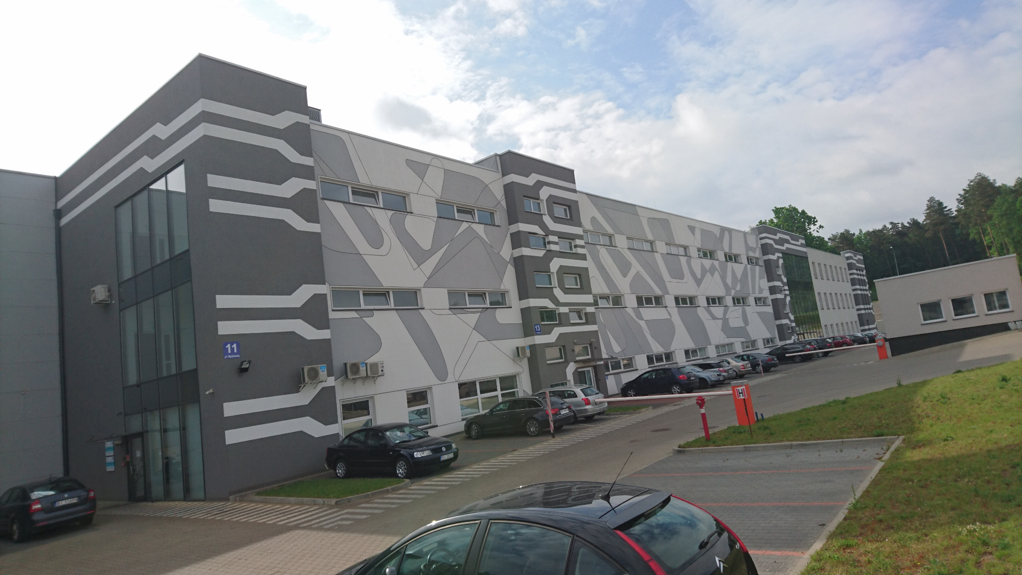 Nowoczesny budynek biurowy z szarą elewacją ozdobioną białymi, geometrycznymi wzorami, widoczne okna, klimatyzatory oraz parking z samochodami.