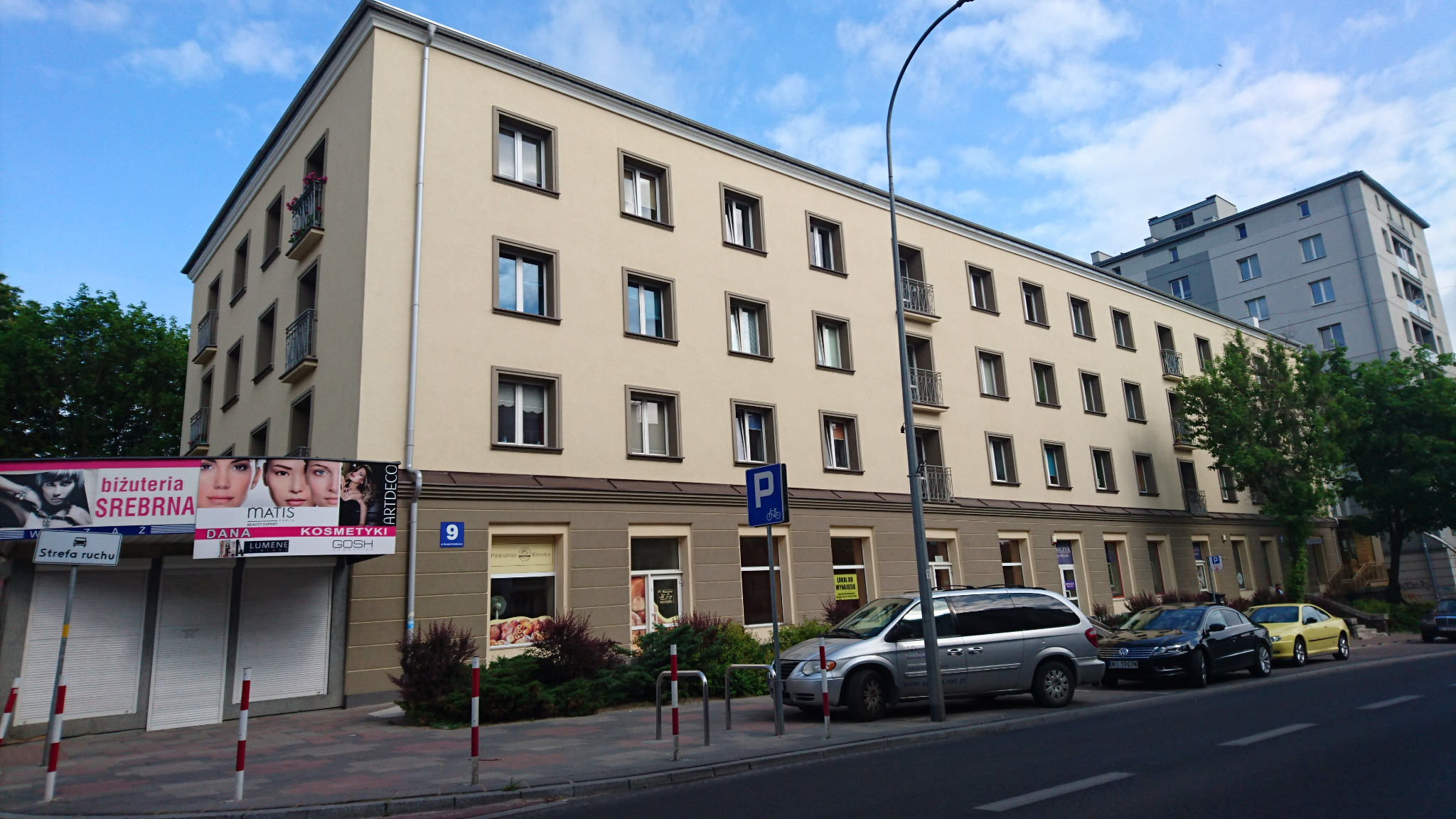Wielopiętrowy budynek mieszkalny z elewacją w odcieniach beżu i brązu, z balkonami i oknami z ciemnymi ramami, widok z poziomu ulicy z zaparkowanymi samochodami i reklamami sklepów na parterze.