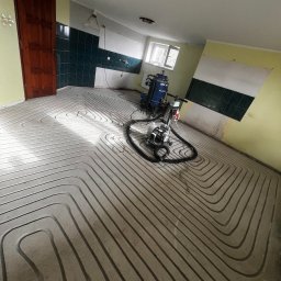 Matusiak Firma Inwestycyjno-Budowlana Krystian Matusiak - Wnętrze w trakcie prac: ułożone rury ogrzewania podłogowego w jastrychu, widoczny sprzęt budowlany i fragment ściany z zielonymi płytkami. Przygotowanie do wylewki.