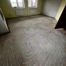 Matusiak Firma Inwestycyjno-Budowlana Krystian Matusiak - Ułożone rury ogrzewania podłogowego w spiralnym wzorze na szarej wylewce, widok z góry. Żółte ściany i okno w tle. Etap prac przed zalaniem posadzki.