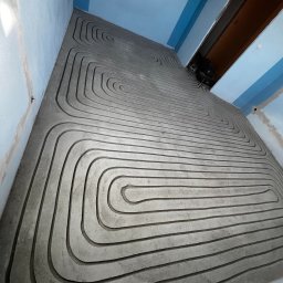 Matusiak Firma Inwestycyjno-Budowlana Krystian Matusiak - Szare rurki ogrzewania podłogowego ułożone w betonowej wylewce, widoczne narzędzia w tle. Ujęcie z góry, ukazujące spiralny wzór instalacji.