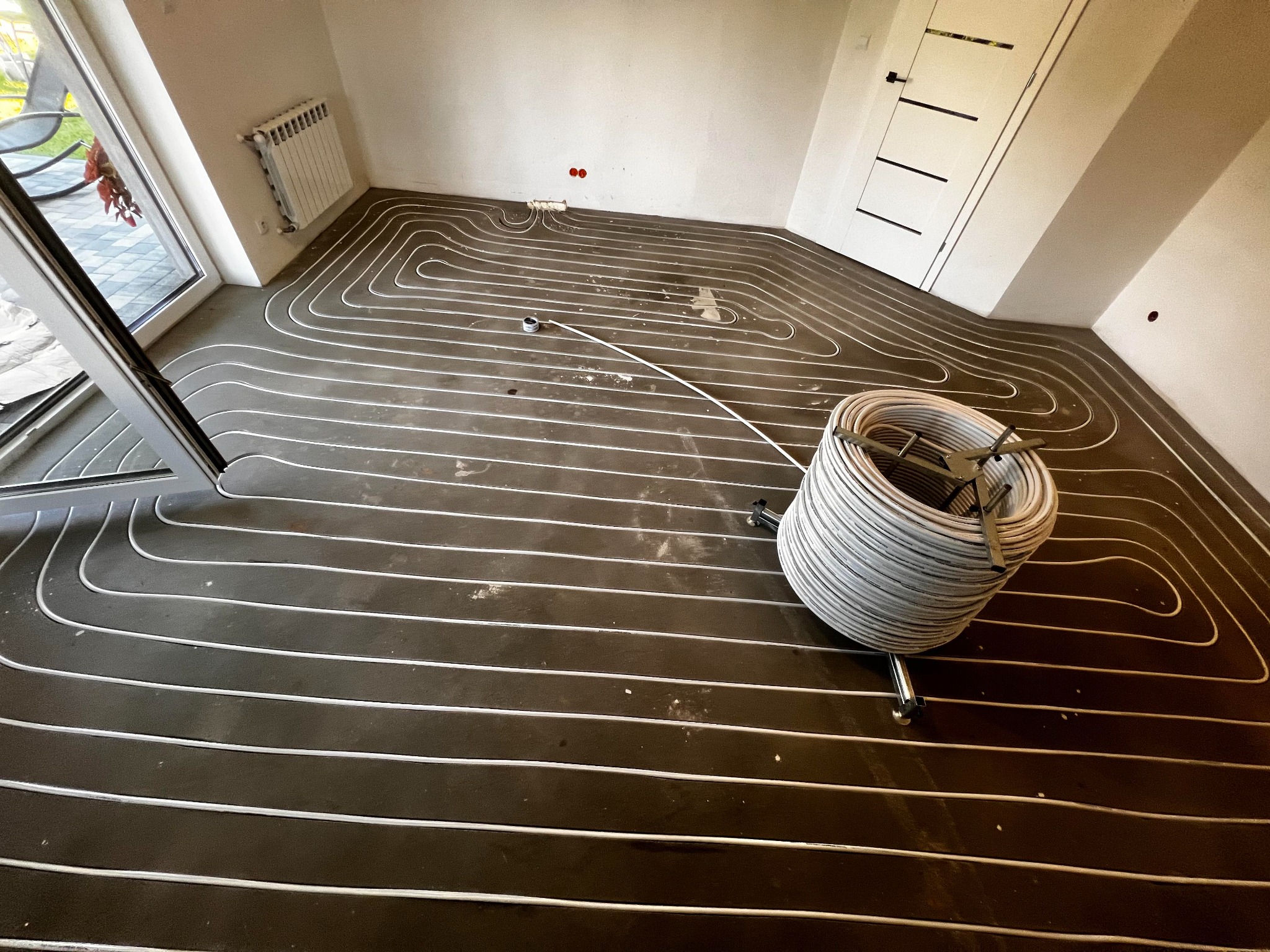 Białe rury ogrzewania podłogowego ułożone spiralnie na ciemnej posadzce w jasnym pomieszczeniu. Widoczny zwój rur i fragment okna.
