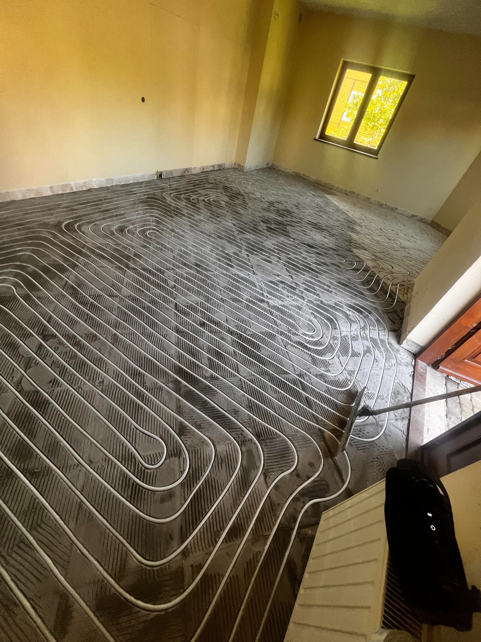 Białe rurki ogrzewania podłogowego ułożone spiralnie na szarej wylewce w jasnym pokoju z oknem. Widok z góry na etap prac przed zalaniem posadzki.
