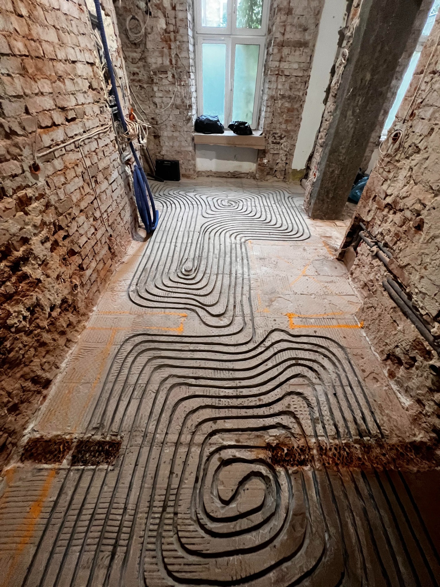 Instalacja ogrzewania podłogowego w surowym wnętrzu z ceglanymi ścianami. Widoczne rury ułożone w spiralny wzór na przygotowanym podłożu. W tle okno i elementy instalacji.