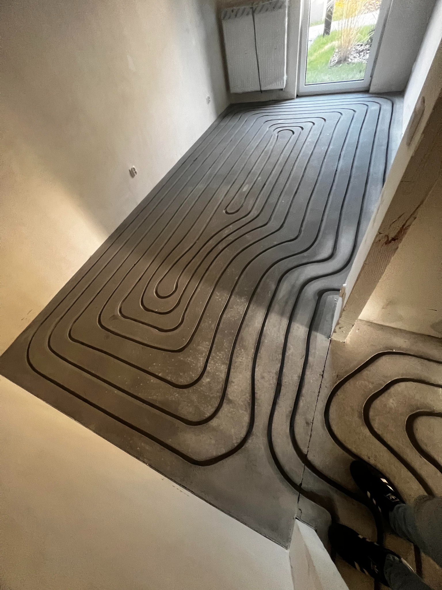 Szare rury ogrzewania podłogowego ułożone spiralnie na wylewce w surowym wnętrzu. Widoczne fragmenty ścian i okno z widokiem na zieleń. Narożnik pomieszczenia.