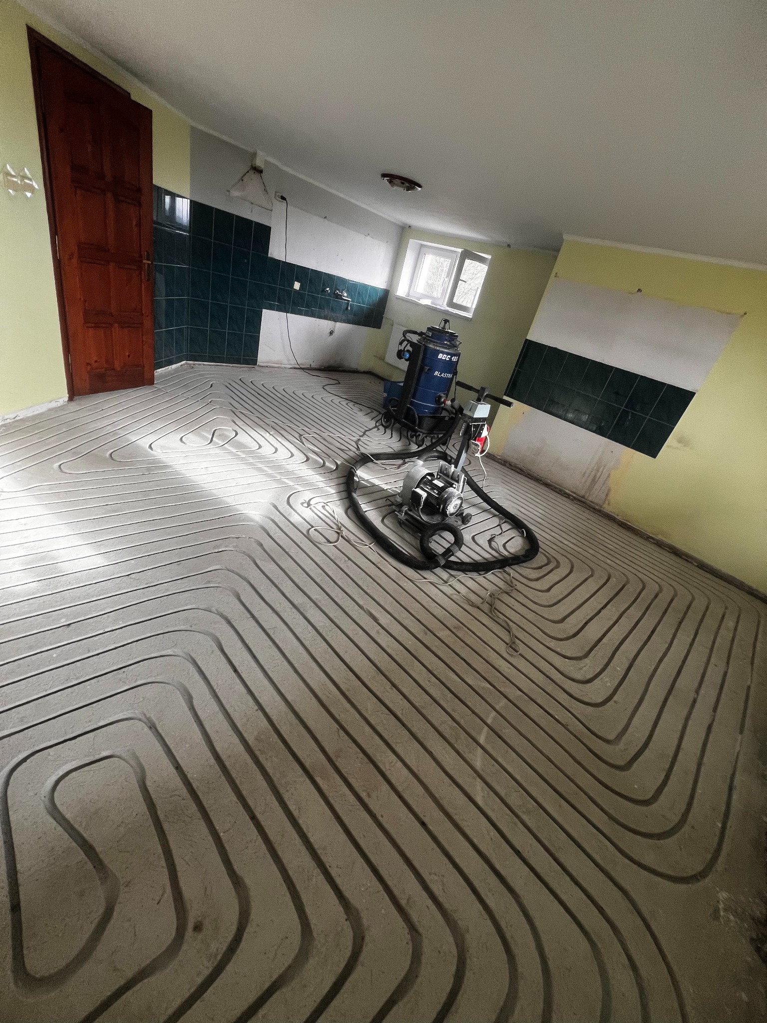 Wnętrze w trakcie prac: ułożone rury ogrzewania podłogowego w jastrychu, widoczny sprzęt budowlany i fragment ściany z zielonymi płytkami. Przygotowanie do wylewki.