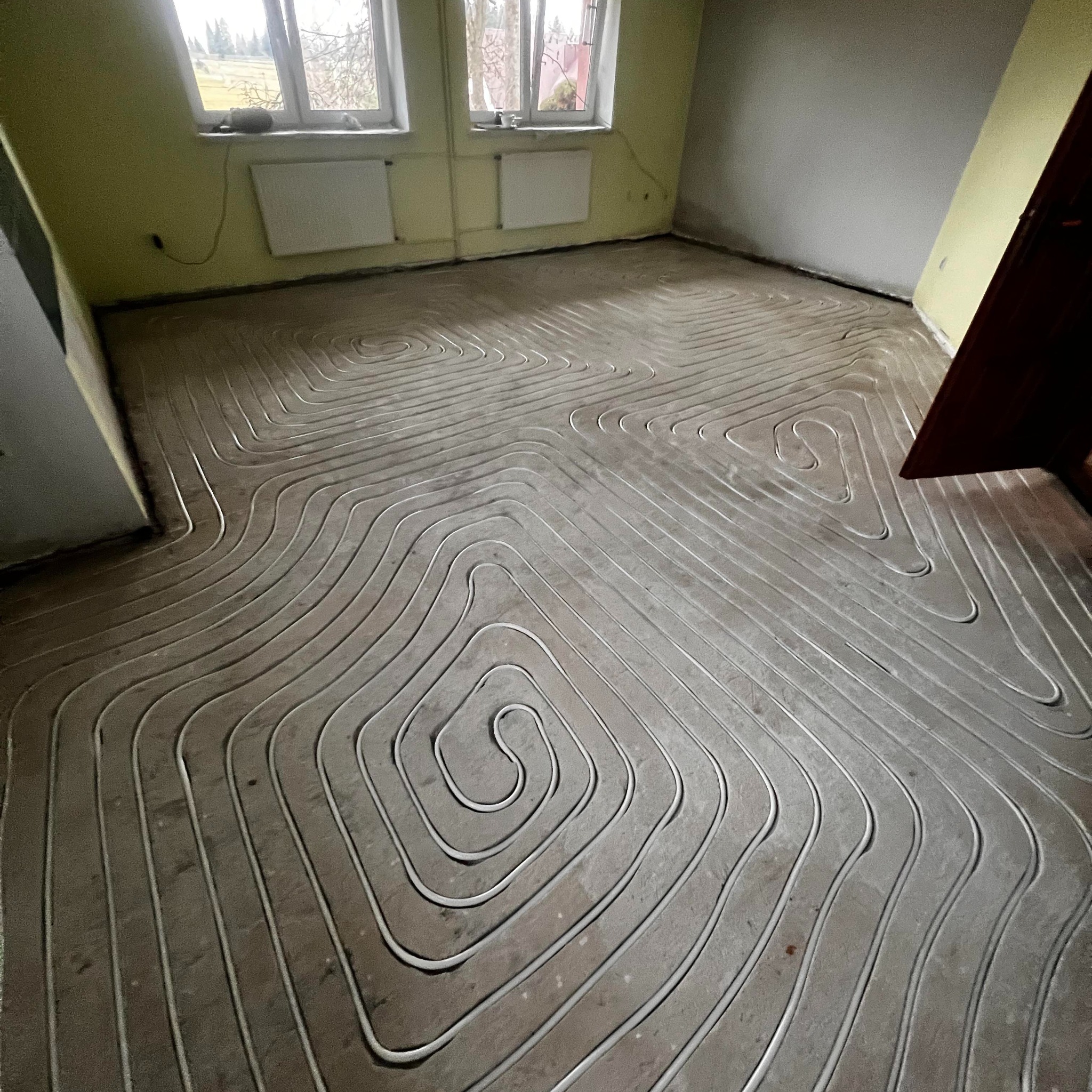 Ułożone rury ogrzewania podłogowego w spiralnym wzorze na szarej wylewce, widok z góry. Żółte ściany i okno w tle. Etap prac przed zalaniem posadzki.