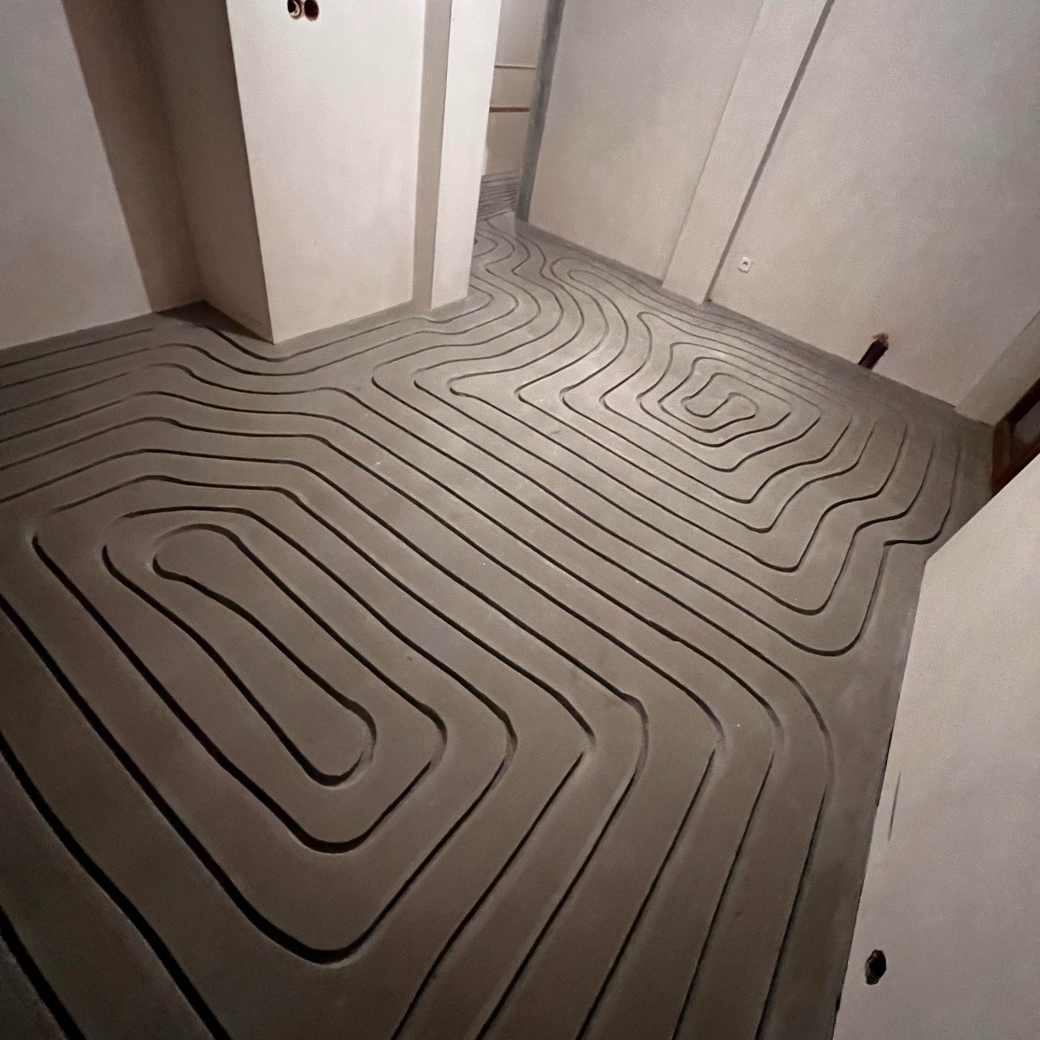 Szare rurki ogrzewania podłogowego ułożone w zygzak na wylewce w surowym wnętrzu. Widoczne ściany w trakcie prac wykończeniowych i wystające kable.