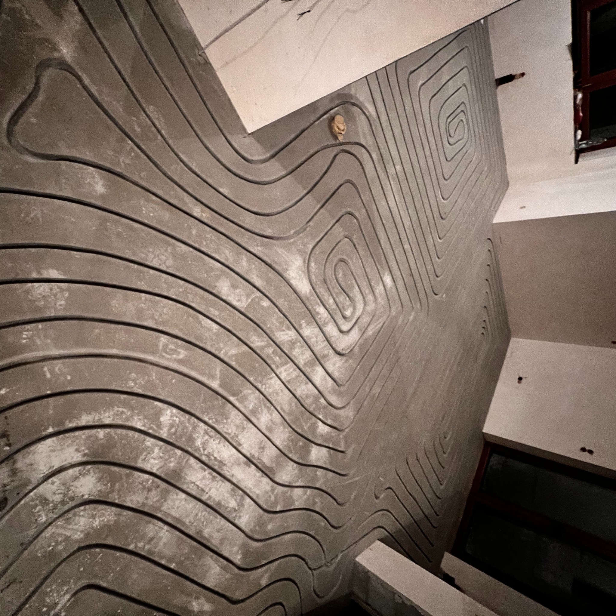 Szare rury ogrzewania podłogowego ułożone spiralnie na wylewce, widoczne okno i fragment sufitu. Ujęcie z góry, ukośne, podkreślające wzór instalacji.