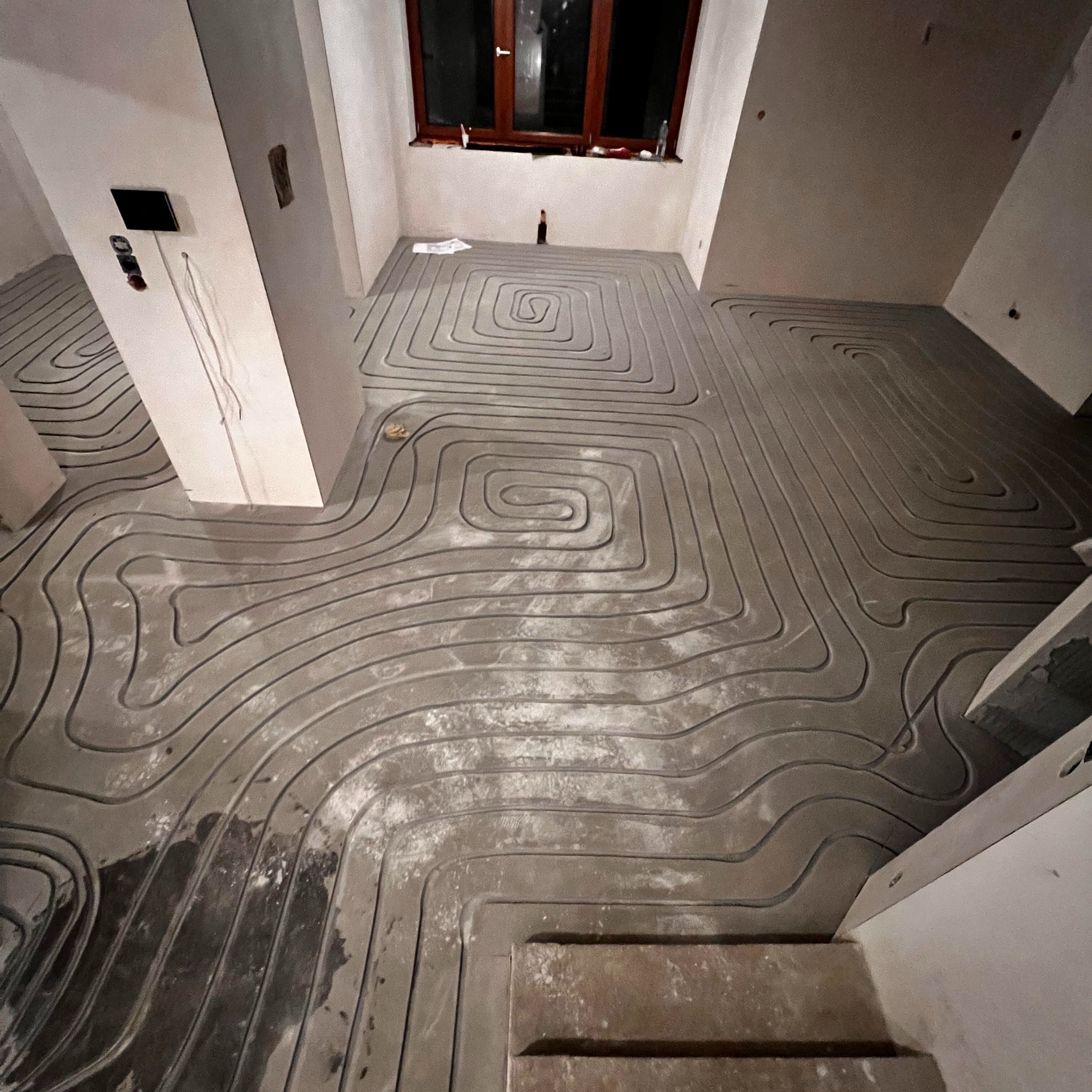 Szare rury ogrzewania podłogowego ułożone spiralnie na betonowej wylewce w surowym wnętrzu. Widoczne okno i fragment ściany z instalacją elektryczną.