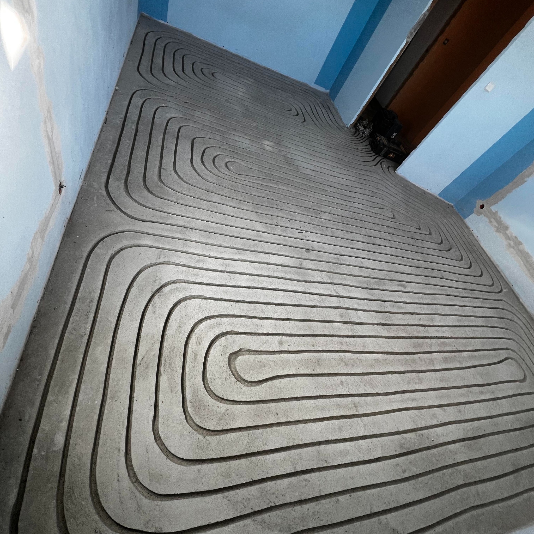 Szare rurki ogrzewania podłogowego ułożone w betonowej wylewce, widoczne narzędzia w tle. Ujęcie z góry, ukazujące spiralny wzór instalacji.