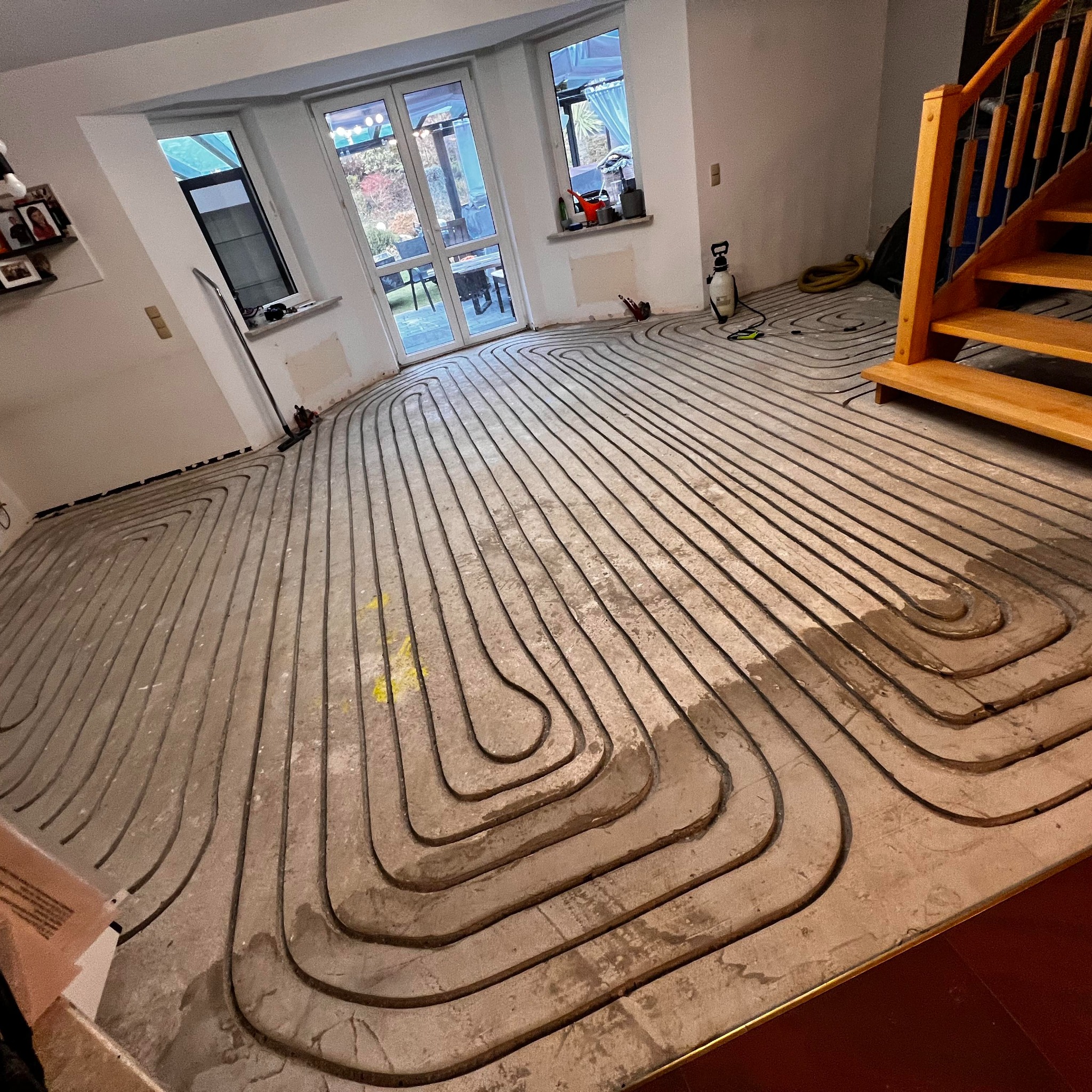 Rozłożona instalacja ogrzewania podłogowego w jasnym pomieszczeniu z oknami i drewnianymi schodami. Widoczne rury ułożone w charakterystyczny wzór na szarej powierzchni.